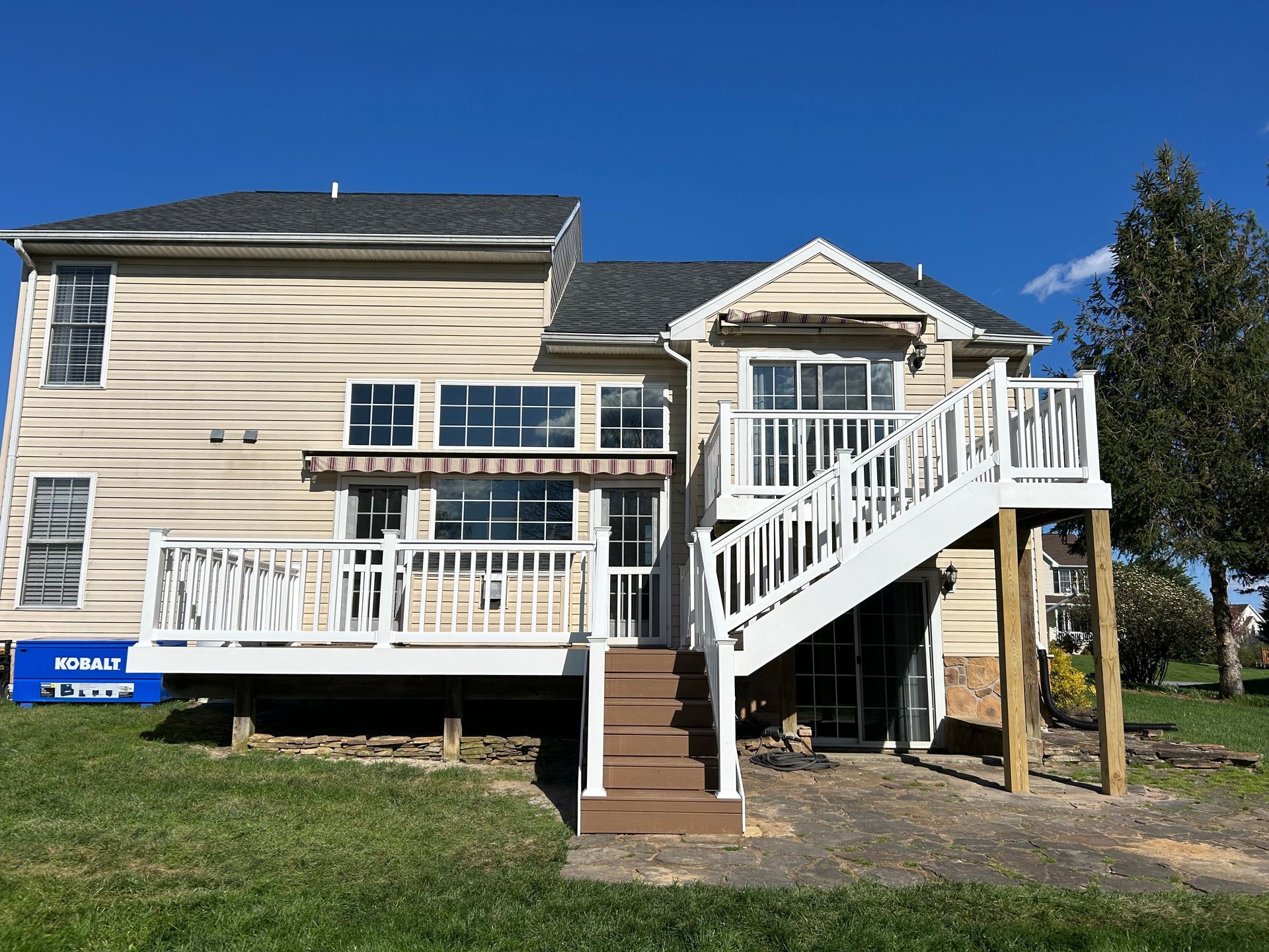 A tan two-story house with a white wooden deck, stairs, and retractable awning under a bright blue sky.