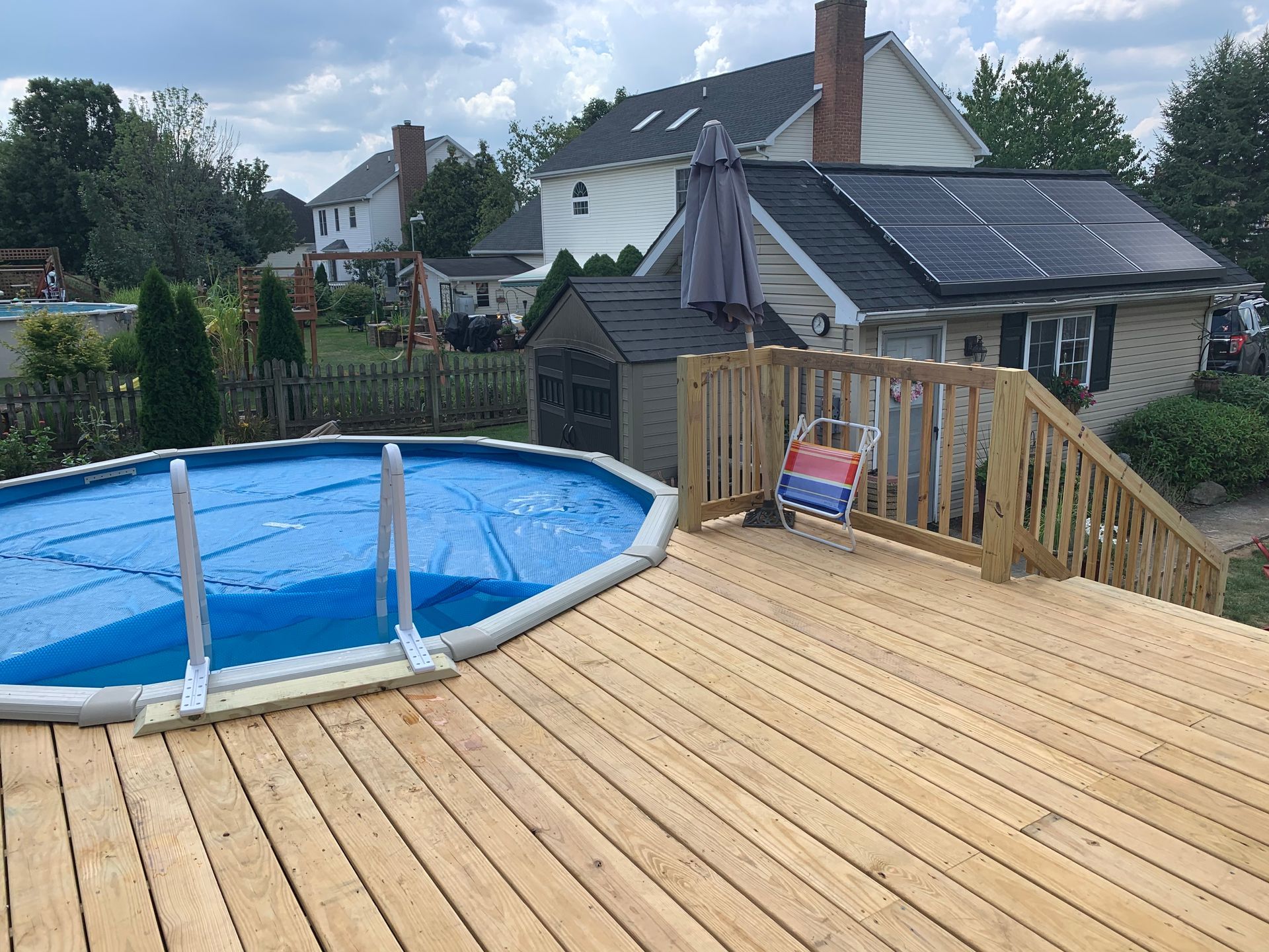 An above-ground swimming pool with a blue cover, set beside a new wooden deck and a house with solar panels.