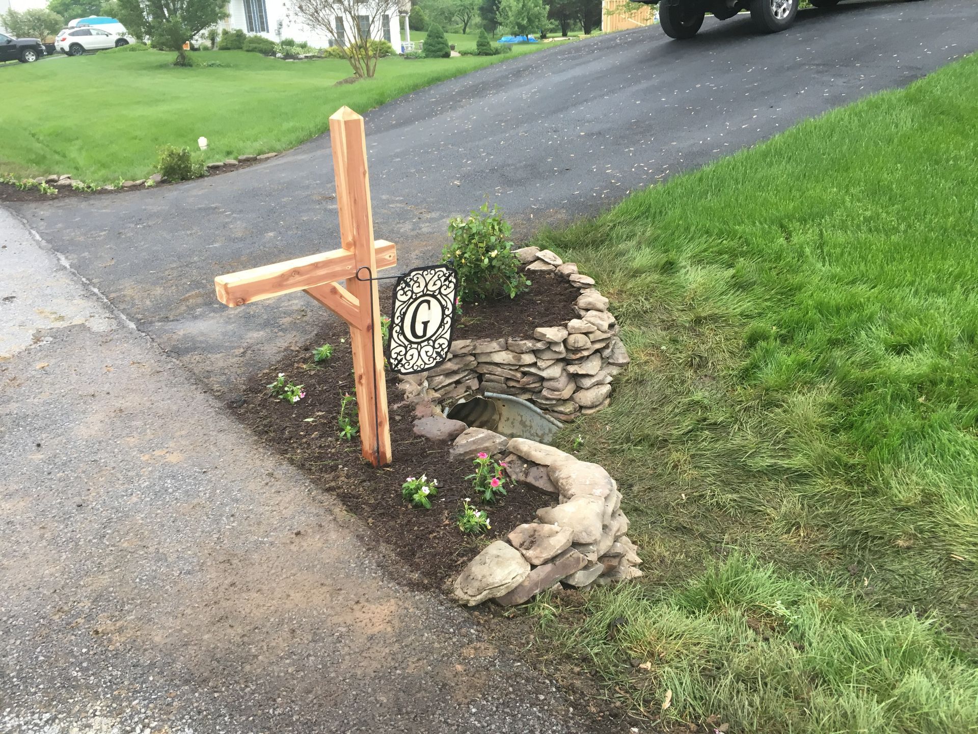 A wooden mailbox post stands in a stone-bordered garden bed at the edge of a driveway, featuring a hanging letter G sign.