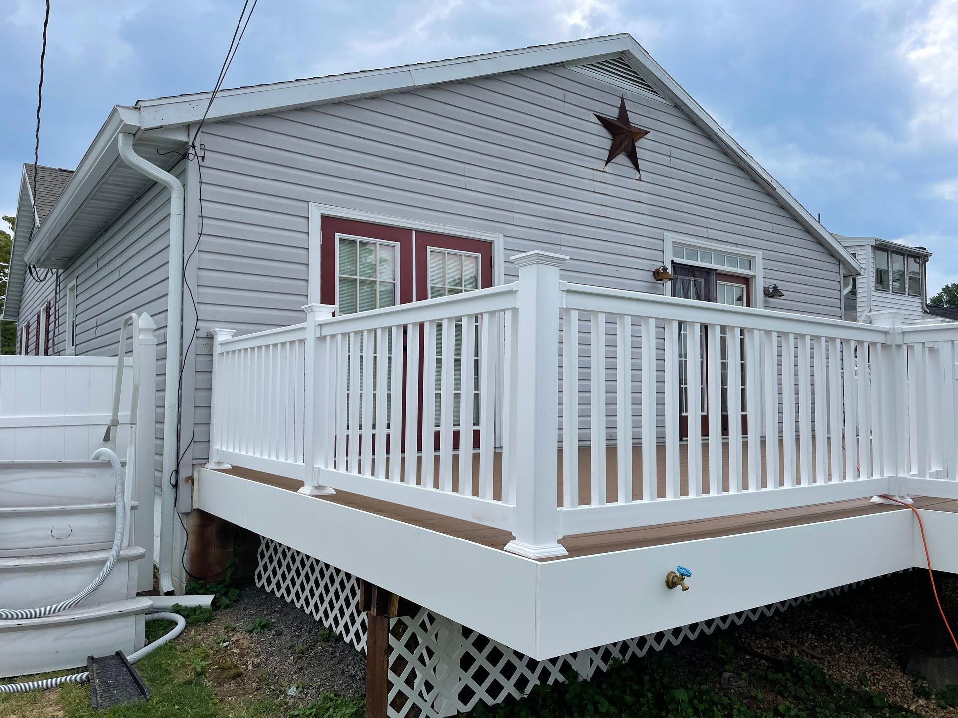 Light gray house with a tan deck and white railing, featuring a red-trimmed door and a decorative star on the gable.