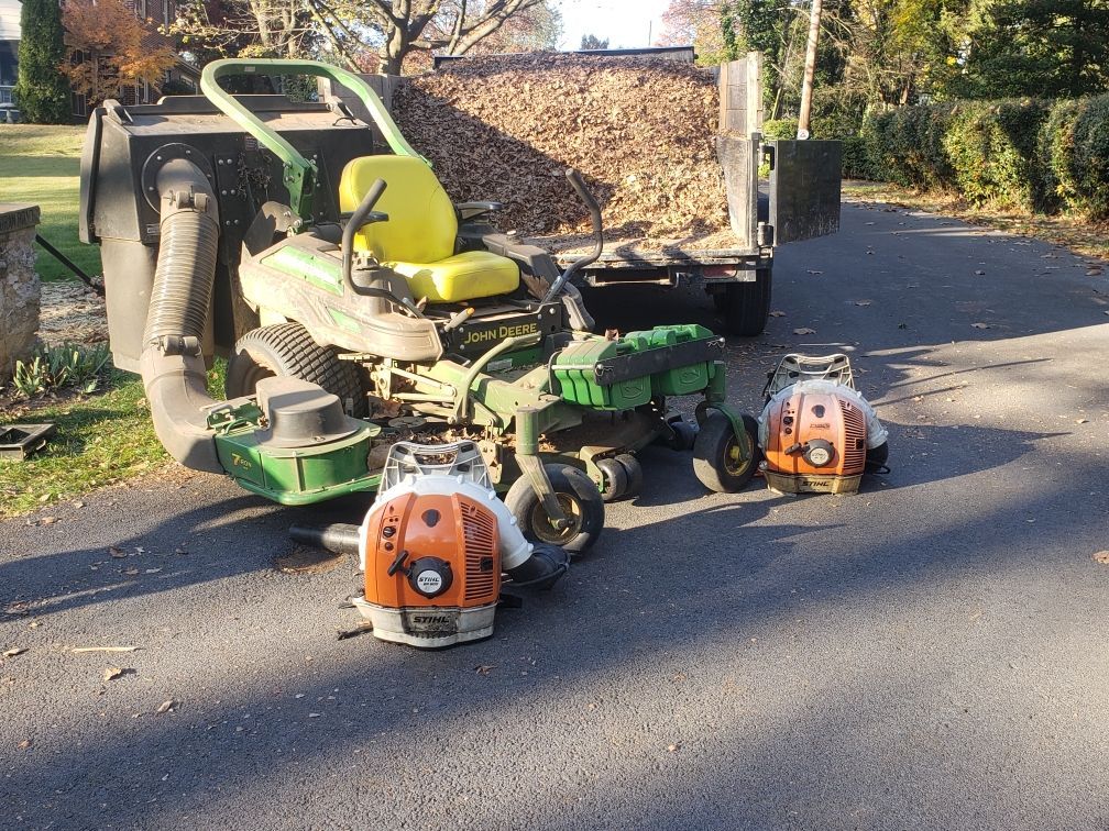A John Deere mower with a leaf collection system and two orange and white leaf blowers parked on a driveway.