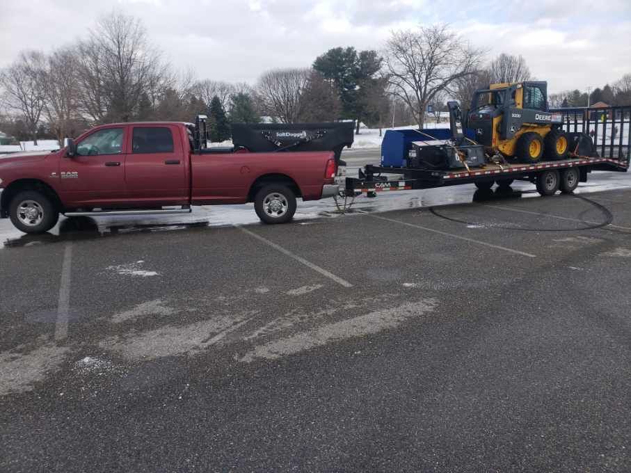 Red pickup truck towing a flatbed trailer with a John Deere skid steer loader in a snowy parking lot.