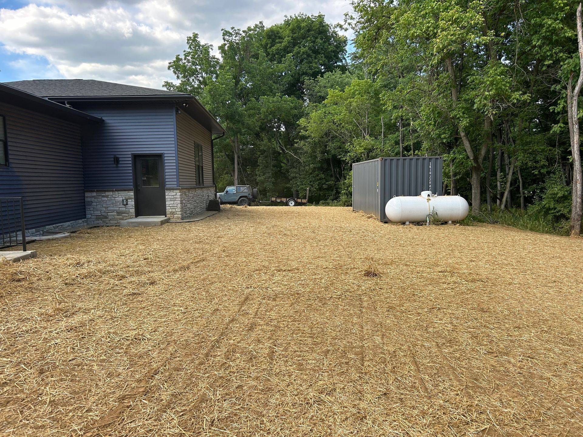 A side view of a house with dark siding and stone accents next to a propane tank, surrounded by a wood chip ground covering.