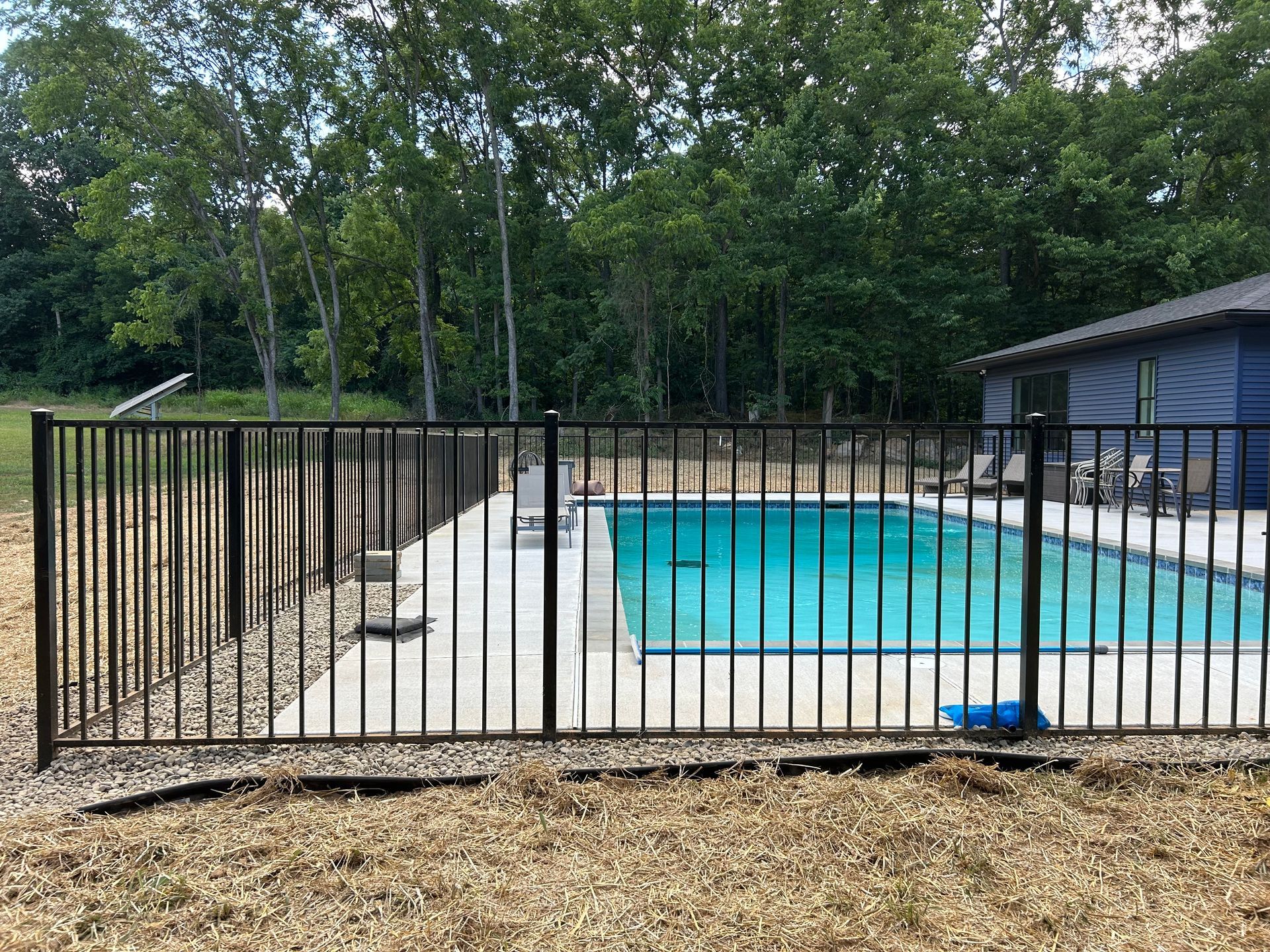 An outdoor swimming pool surrounded by a black metal fence with a house and trees in the background.