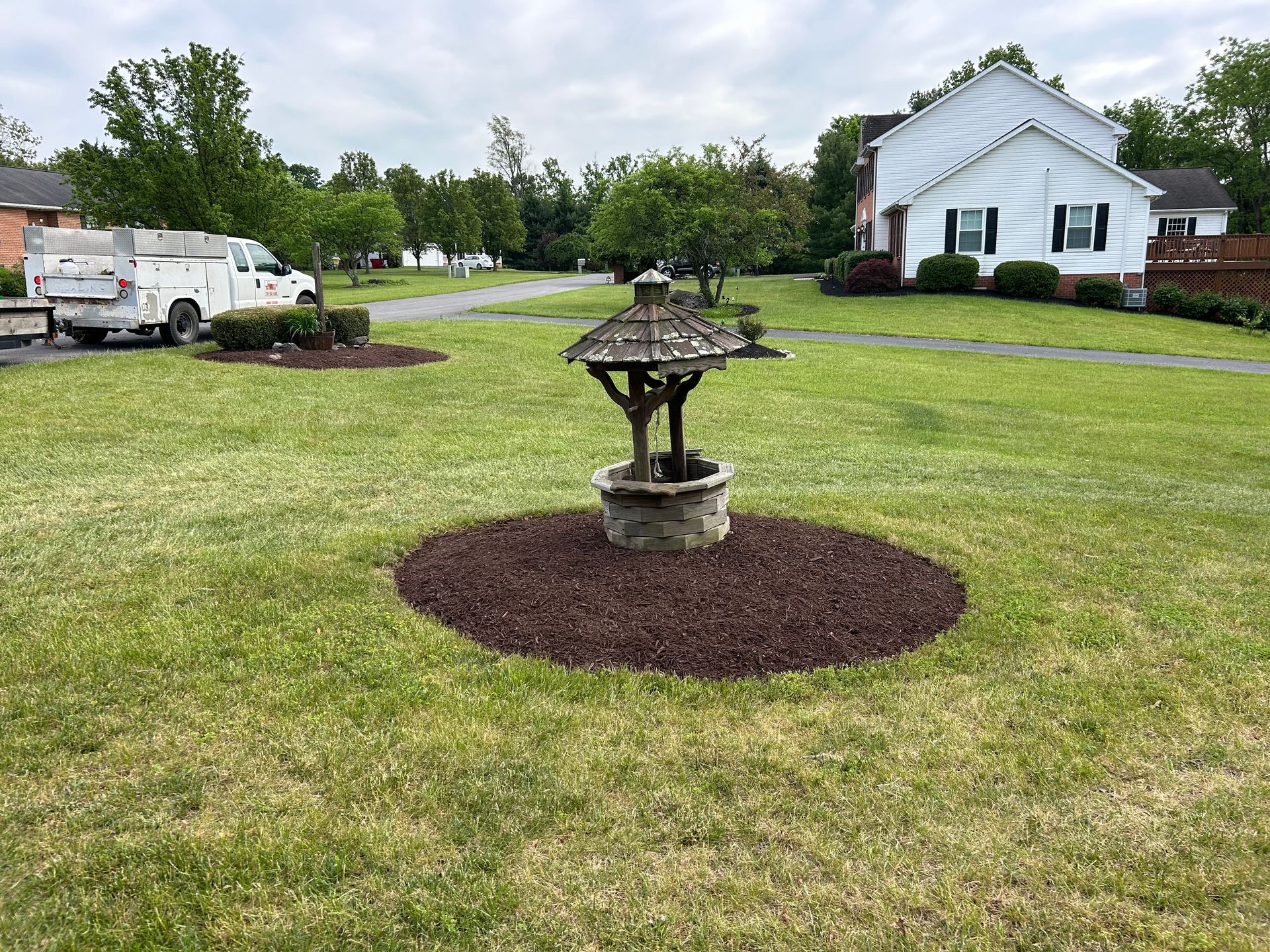 A decorative wooden wishing well stands in the center of a mulch circle on a green lawn, with a house and van nearby.