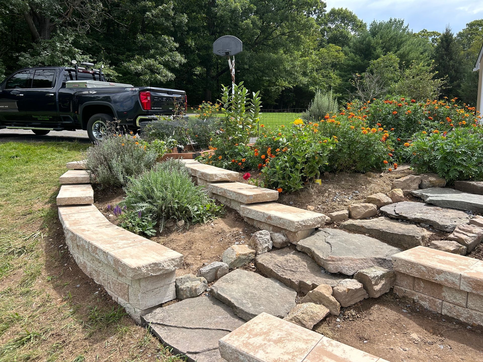 A curved stone retaining wall borders a garden bed with flowers and flagstone steps in a yard with a black truck parked.