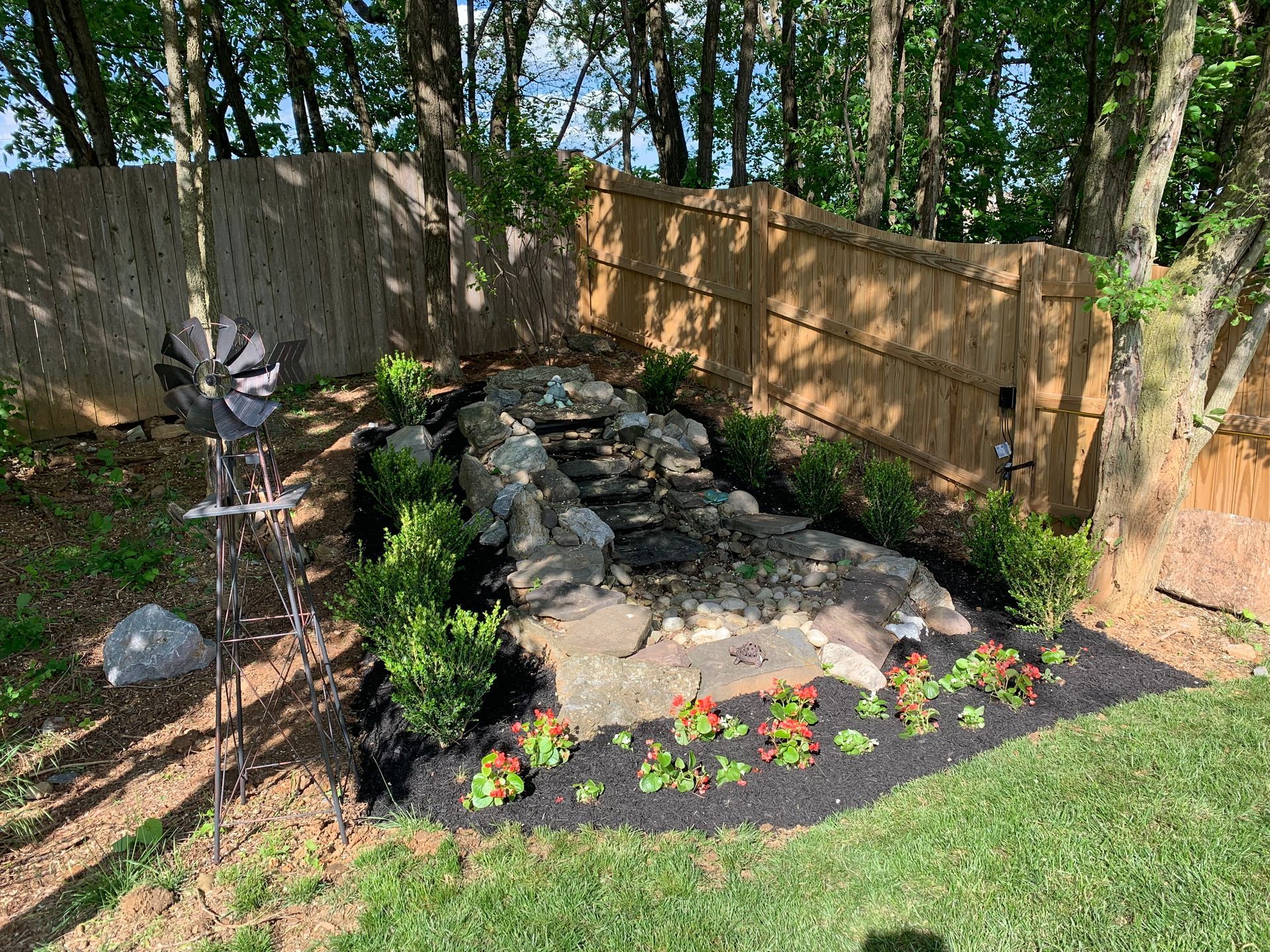 A stone-lined water feature in a backyard garden, surrounded by small green bushes, red flowers, and black mulch.