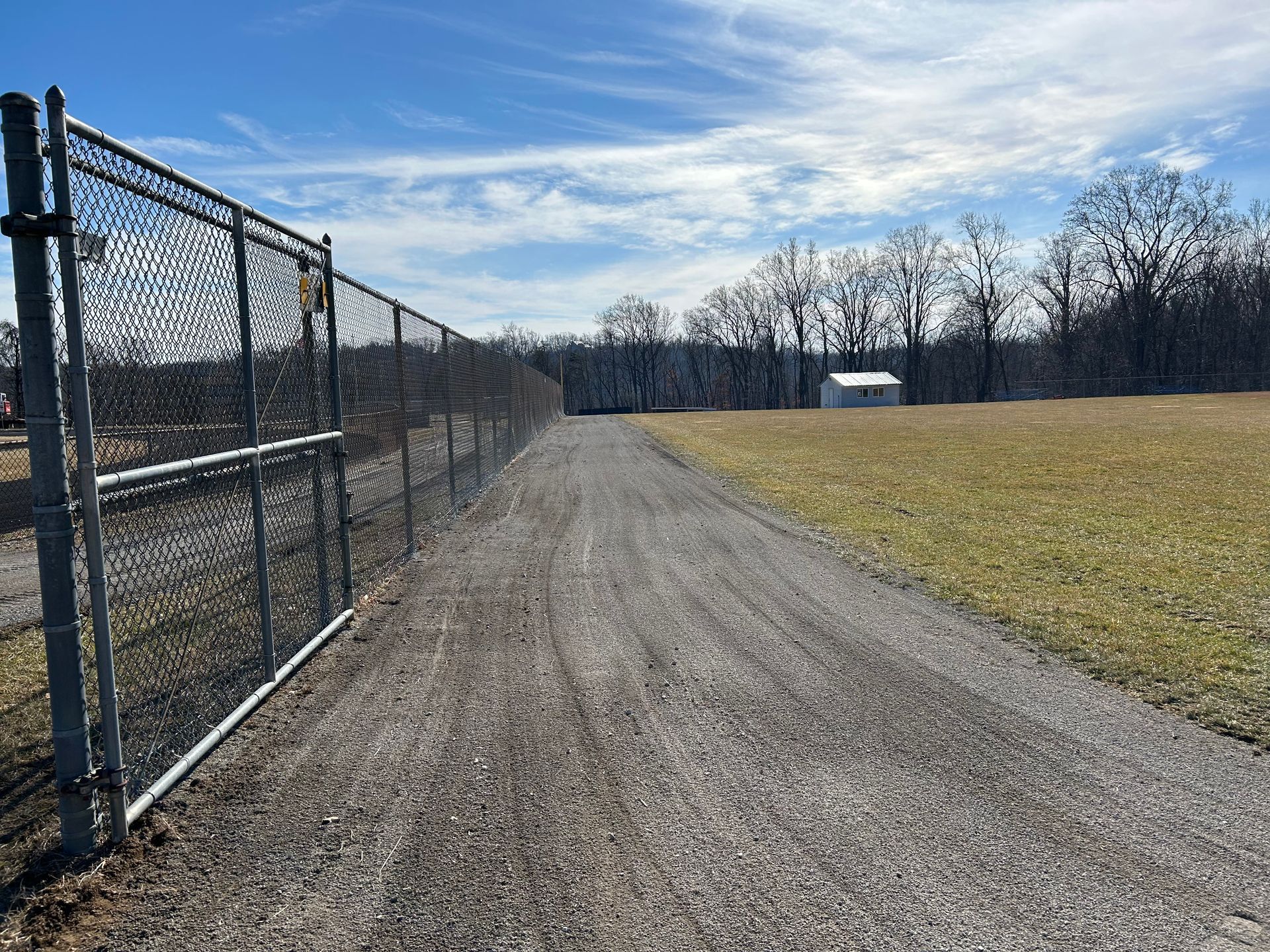 A gravel path stretches between a metal chain-link fence on the left and a field on the right under a bright blue sky.