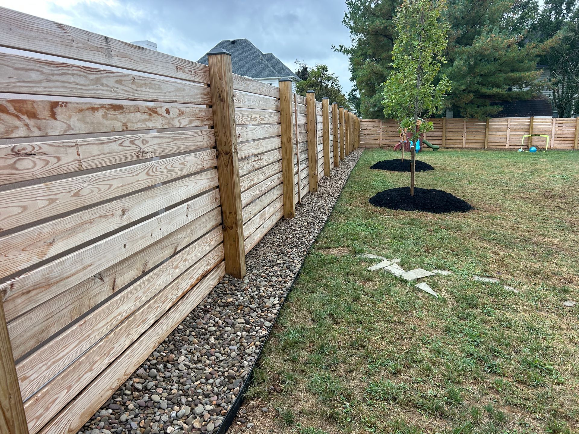 A horizontal wooden plank fence runs along a gravel border in a grassy backyard with two young trees in mulch beds.