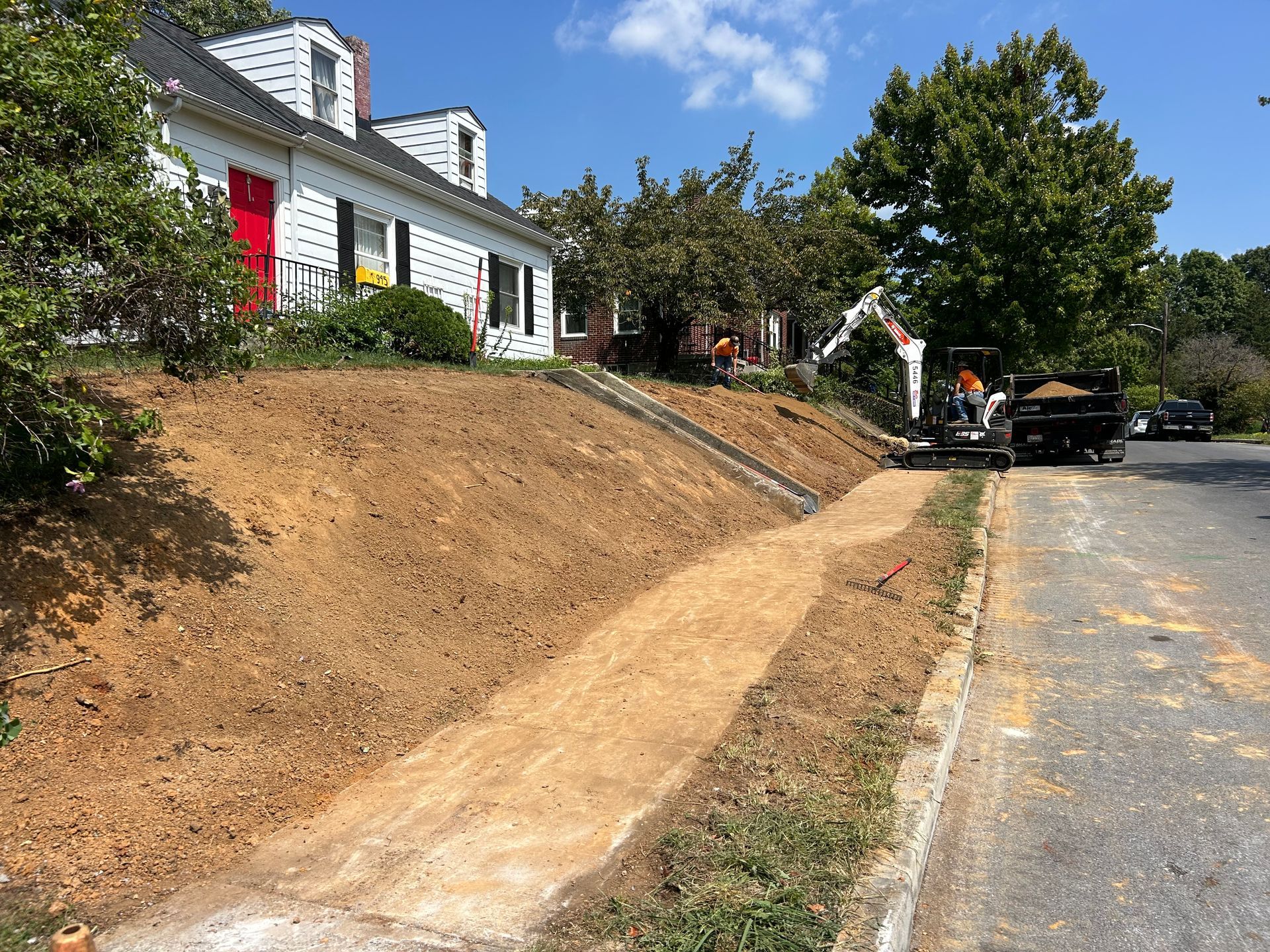 A white house with a red door sits above a freshly excavated dirt slope next to a road where construction is underway.