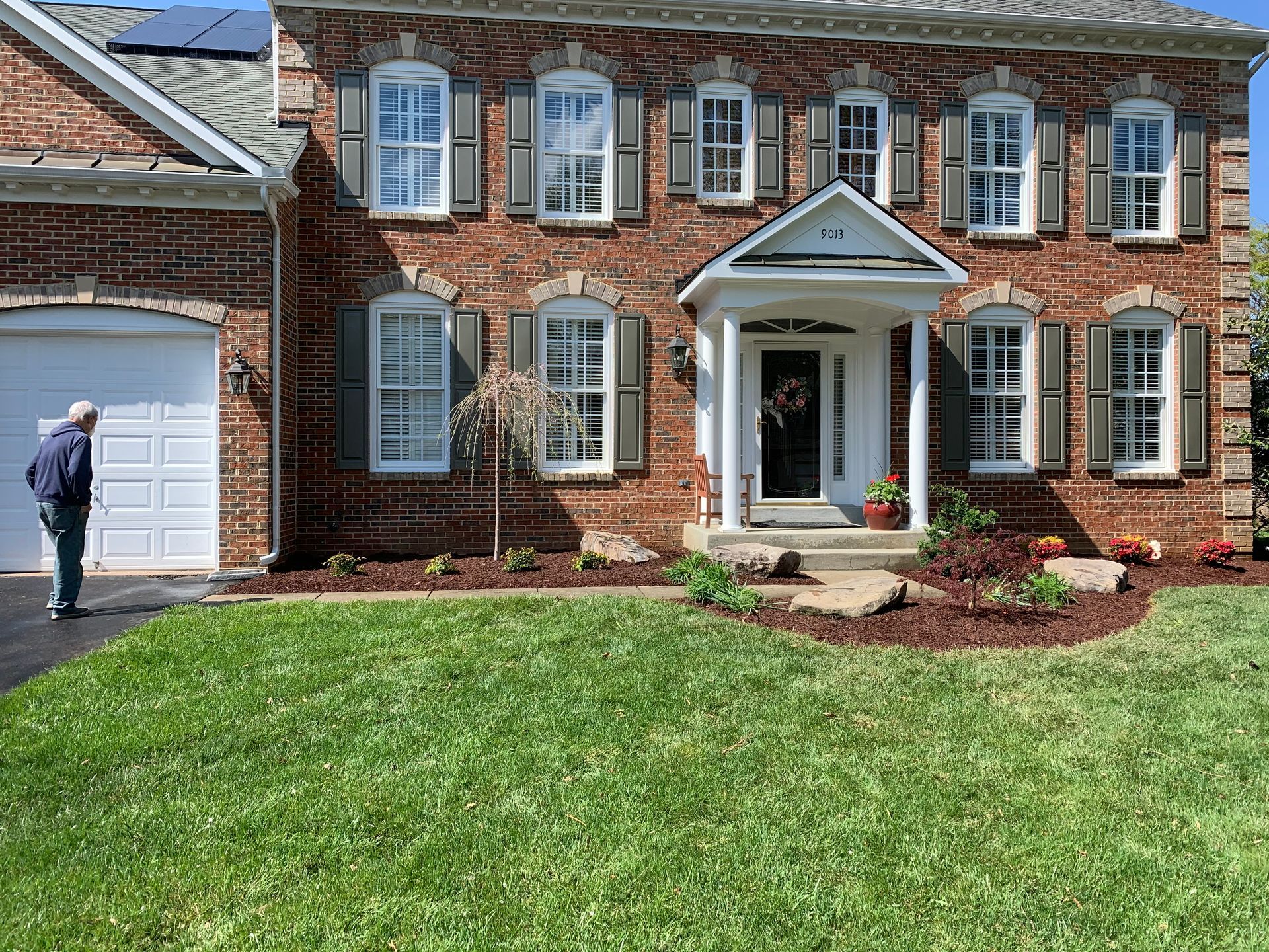 A two-story brick house with a white garage door, front porch, and a person walking on the driveway in a sunny yard.