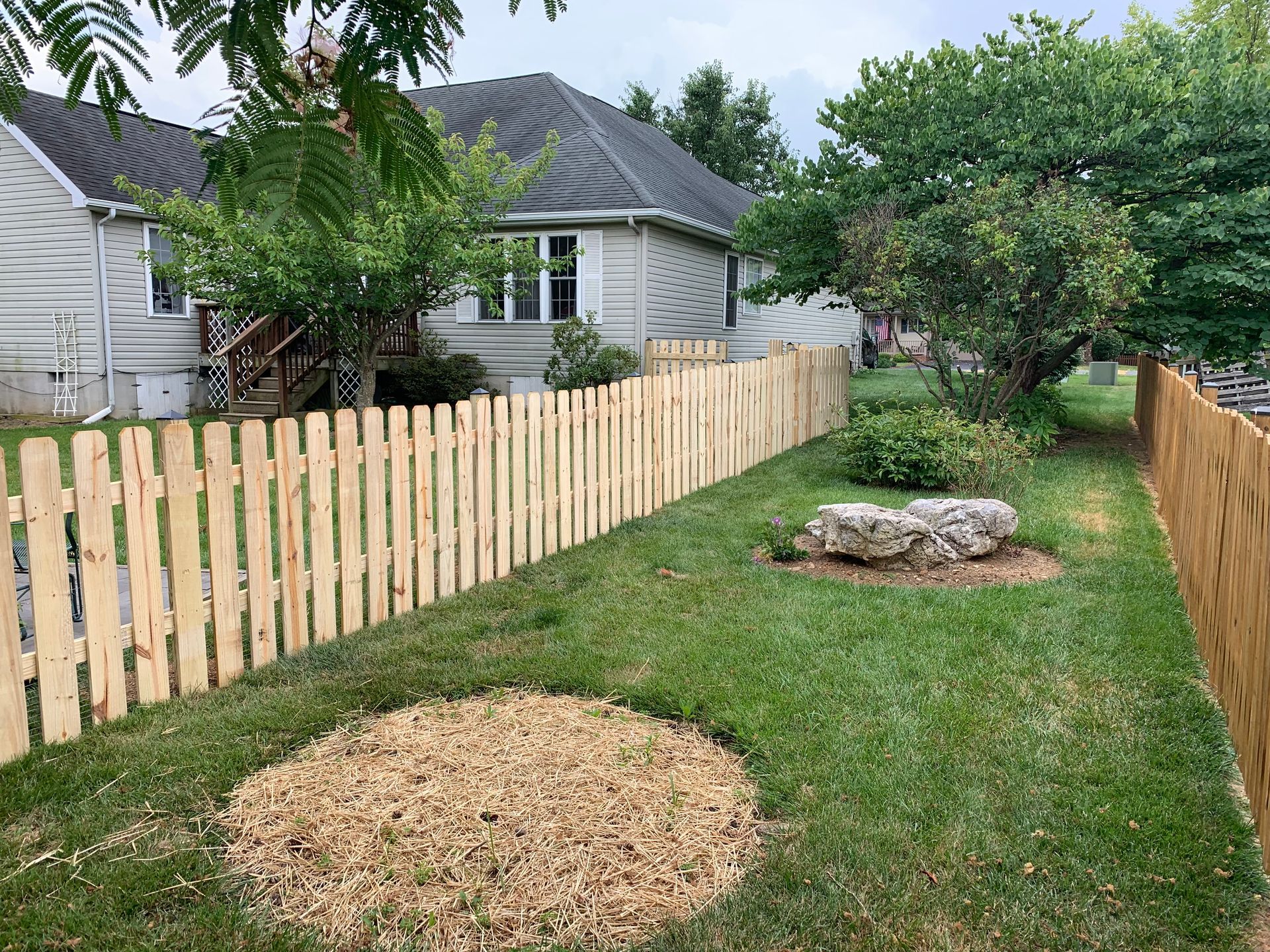 A newly installed light-wood picket fence borders a residential backyard with a mulched circular area and a garden rock.