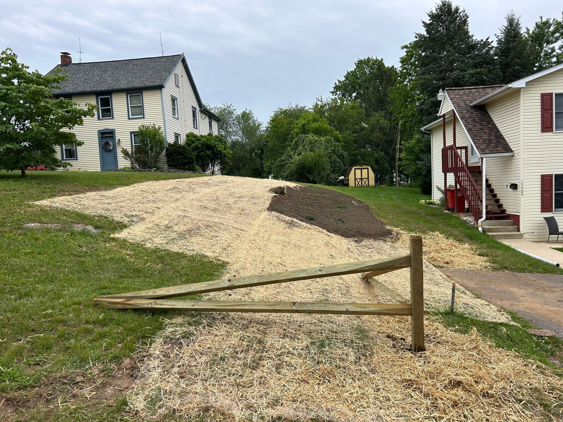 A sloped yard between two houses features a fresh pile of wood chips covering a pathway near a wooden fence post.
