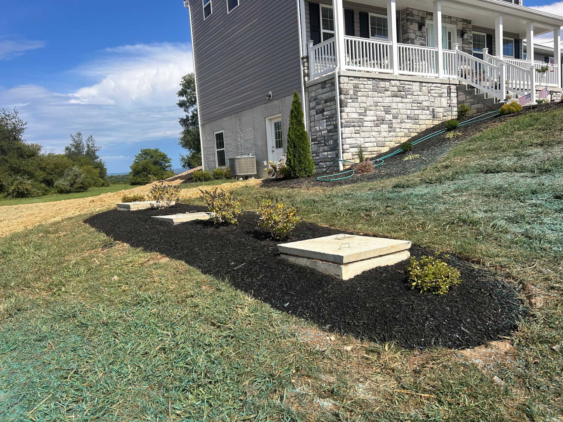 A landscaping bed with black mulch and three concrete septic lids in front of a house with stone siding and a white porch.