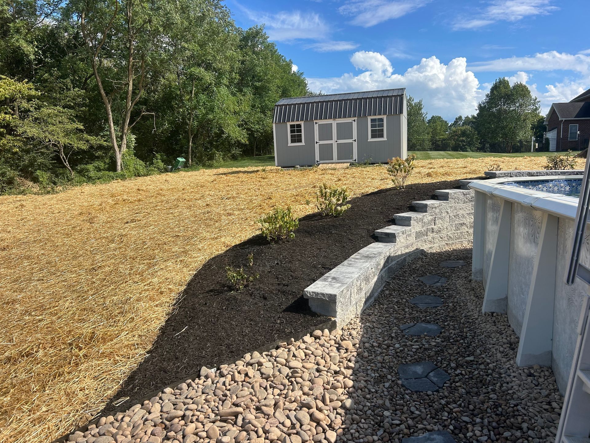 A backyard scene features a gray storage shed, a mulched garden bed with small shrubs, and a gravel path beside a pool.