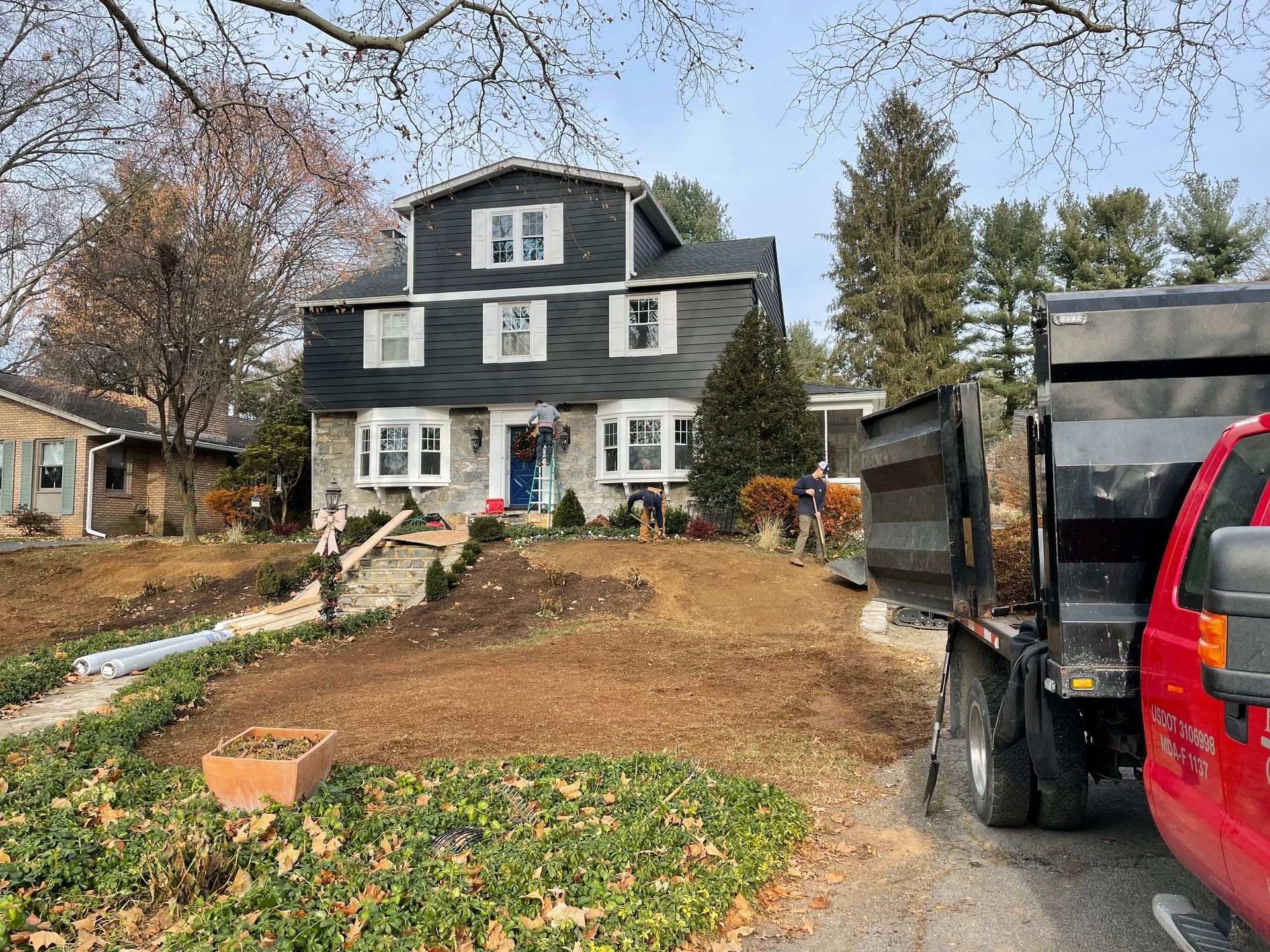 A dark-sided two-story house under renovation with workers in a front yard and a red dump truck parked on the side.