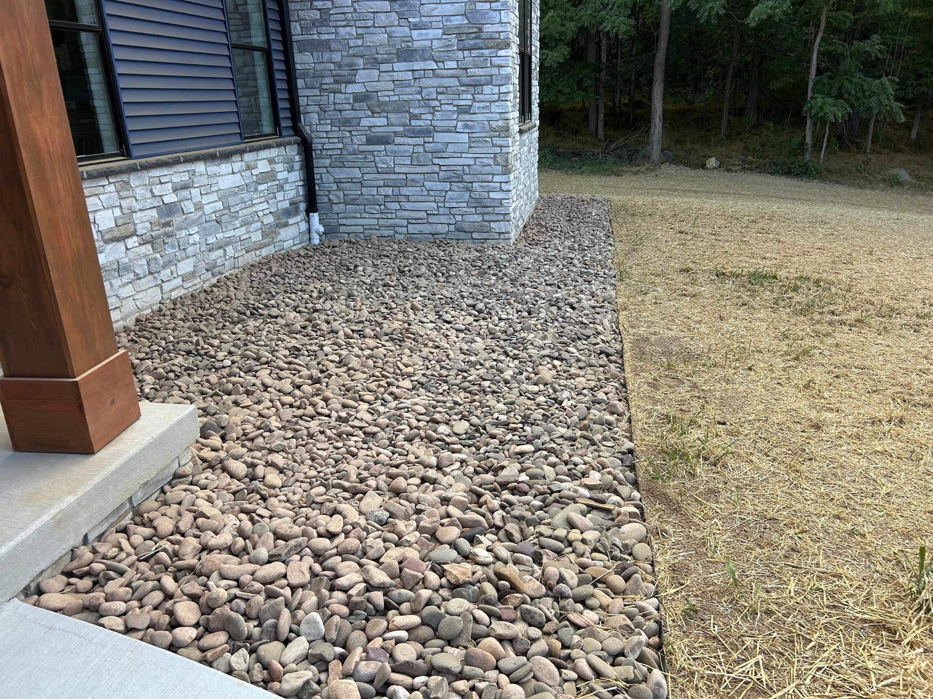 A stone foundation garden bed with brown river rocks next to a building with gray stone siding and a patch of dry grass.