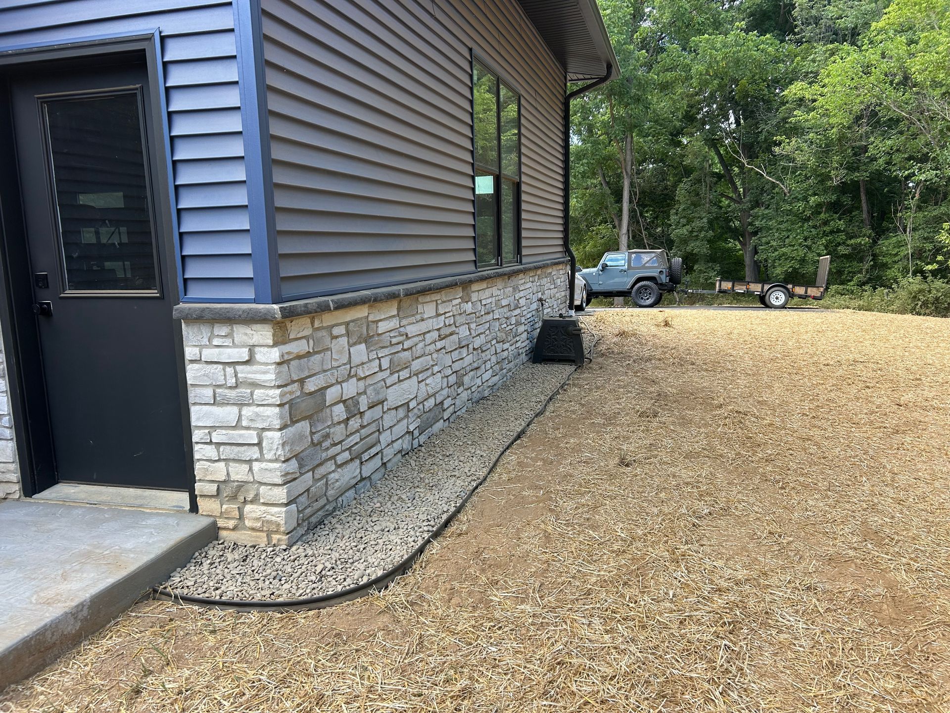 A modern house exterior with gray siding over a stone base, next to a gravel area with a parked car in the background.
