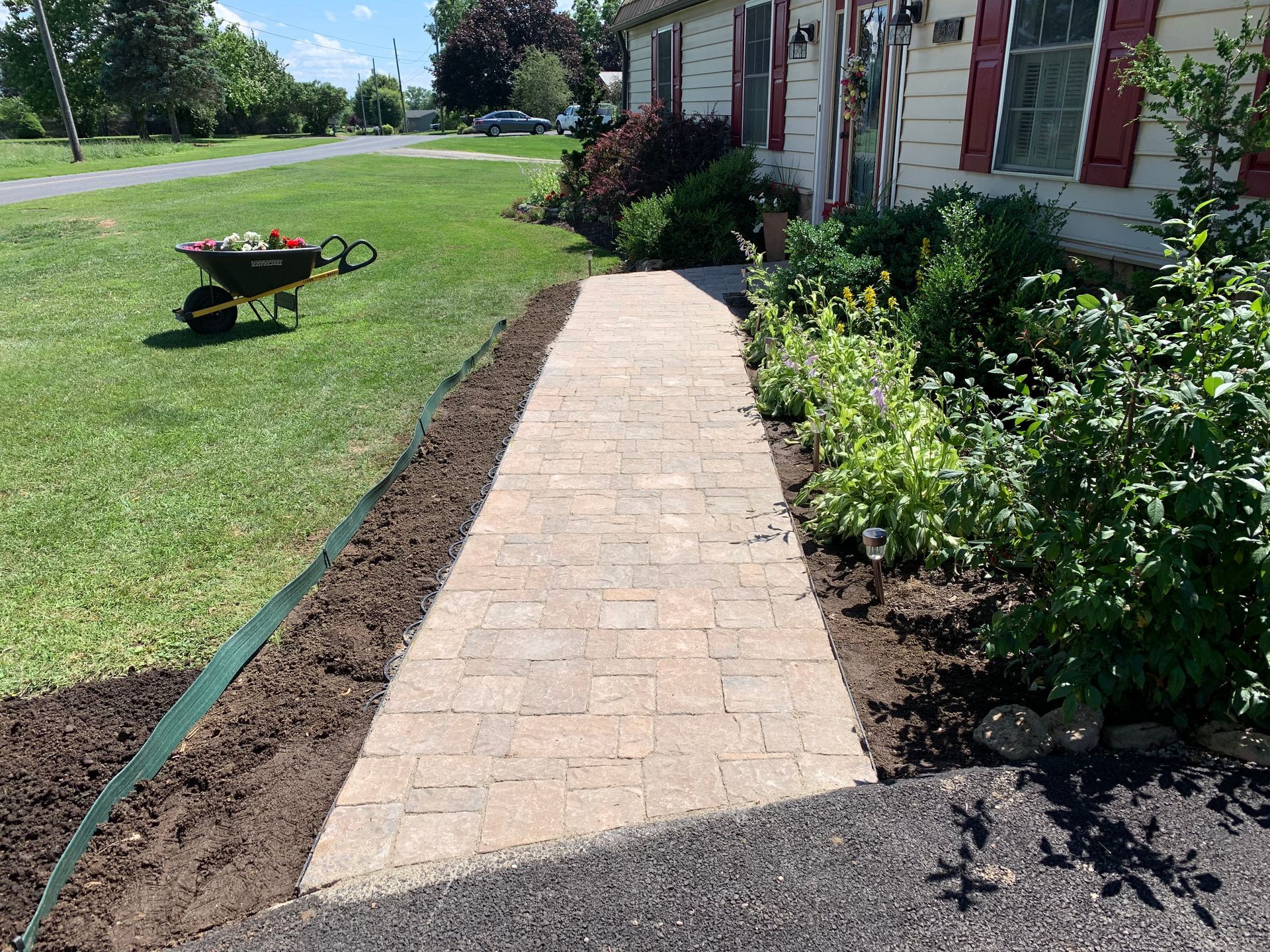 A new paver walkway leading to a house entrance, bordered by soil beds and green landscaping, with a wheelbarrow nearby.