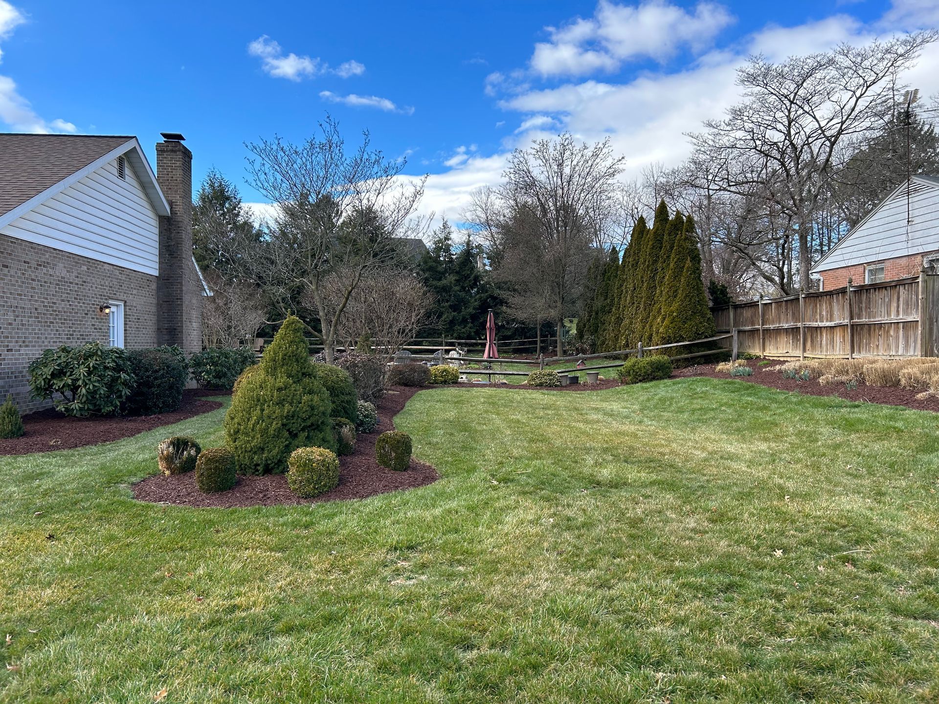 A grassy backyard with a garden bed of shrubs near a brick house, a wooden fence in the back, and trees under a blue sky.