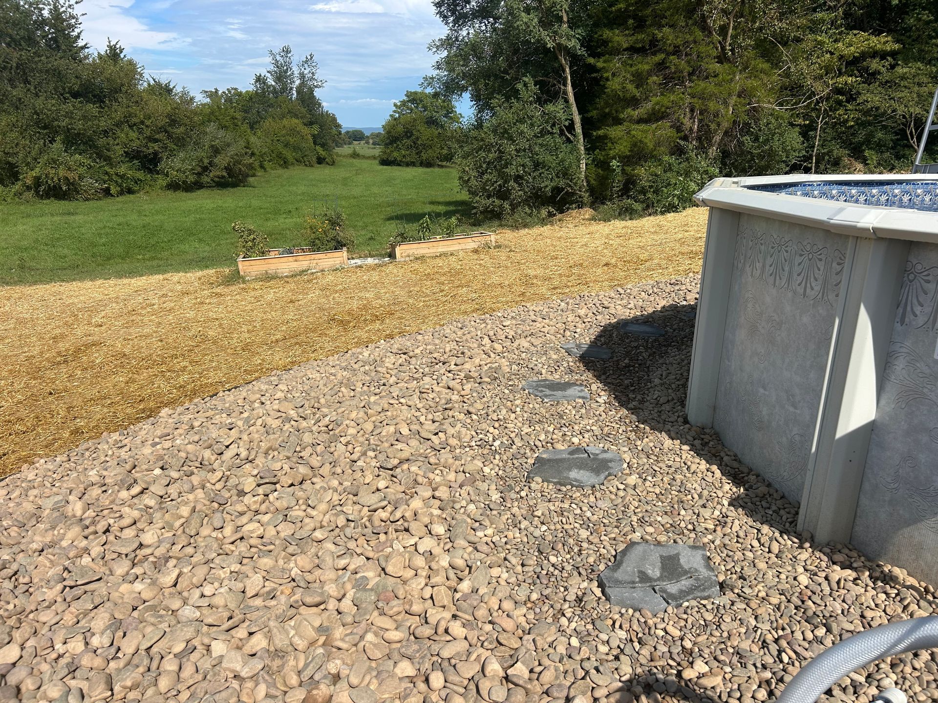 Gravel ground cover surrounding an above-ground swimming pool, adjacent to a grassy lawn and trees under a blue sky.