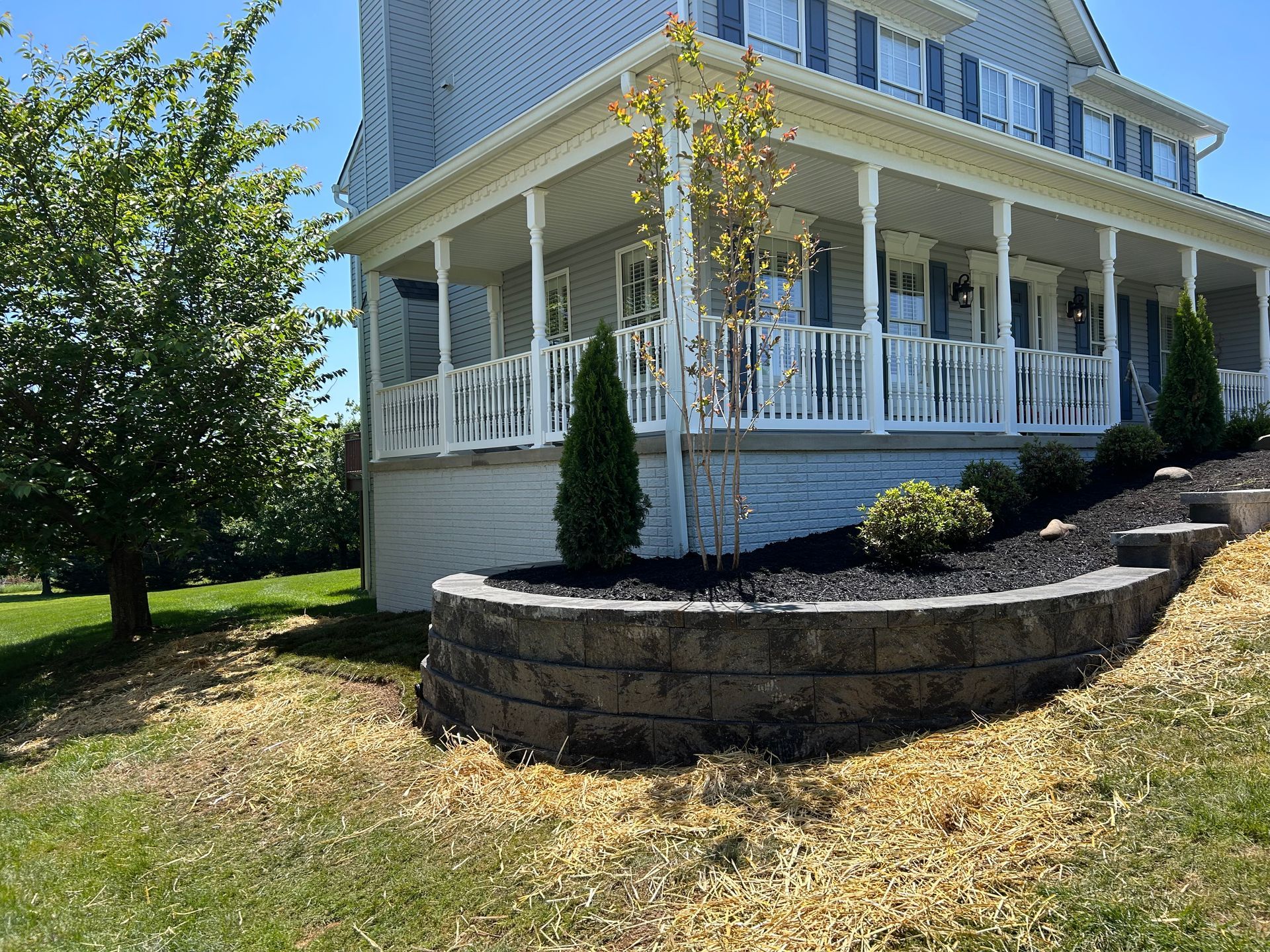 A light blue house with a white front porch features a tiered stone retaining wall and mulch bed with small evergreen trees.