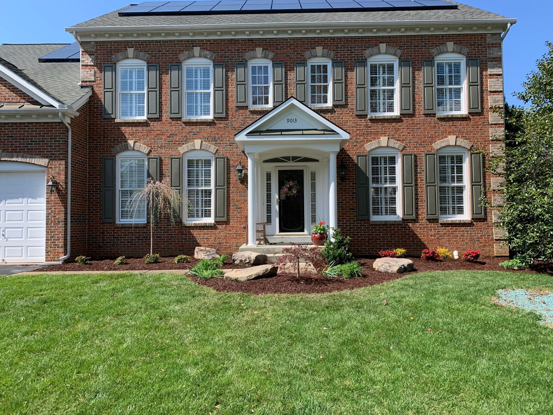 A two-story red brick house with a white front porch entrance, dark shutters, and solar panels on the shingled roof.
