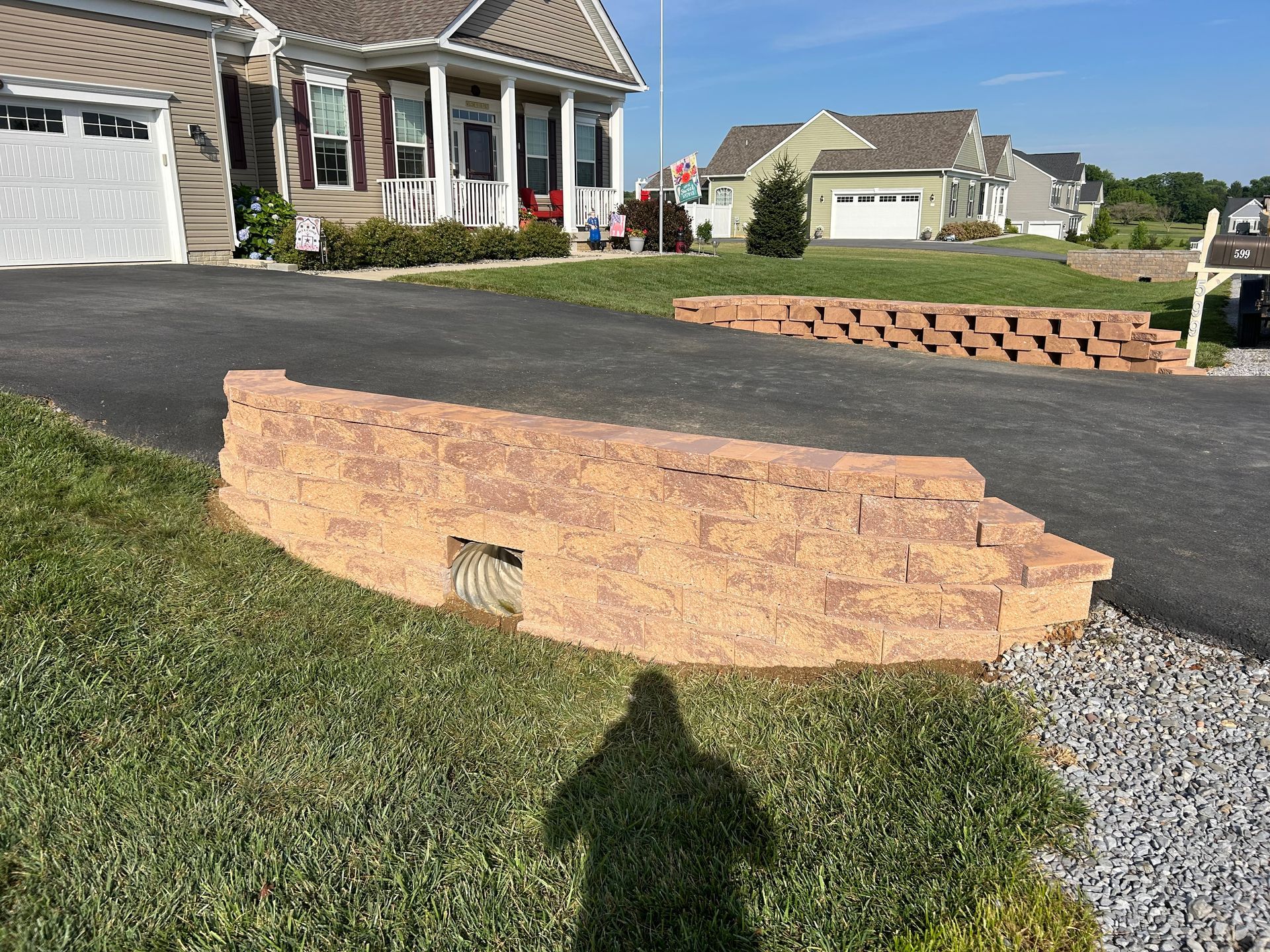 A low, tan stone retaining wall with a drainage pipe built into the base sits beside an asphalt driveway near a house.