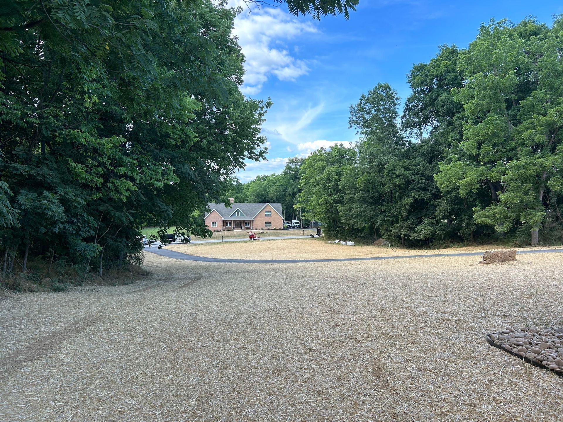 A long gravel driveway leads through a wooded area toward a house in the distance under a blue sky with light clouds.