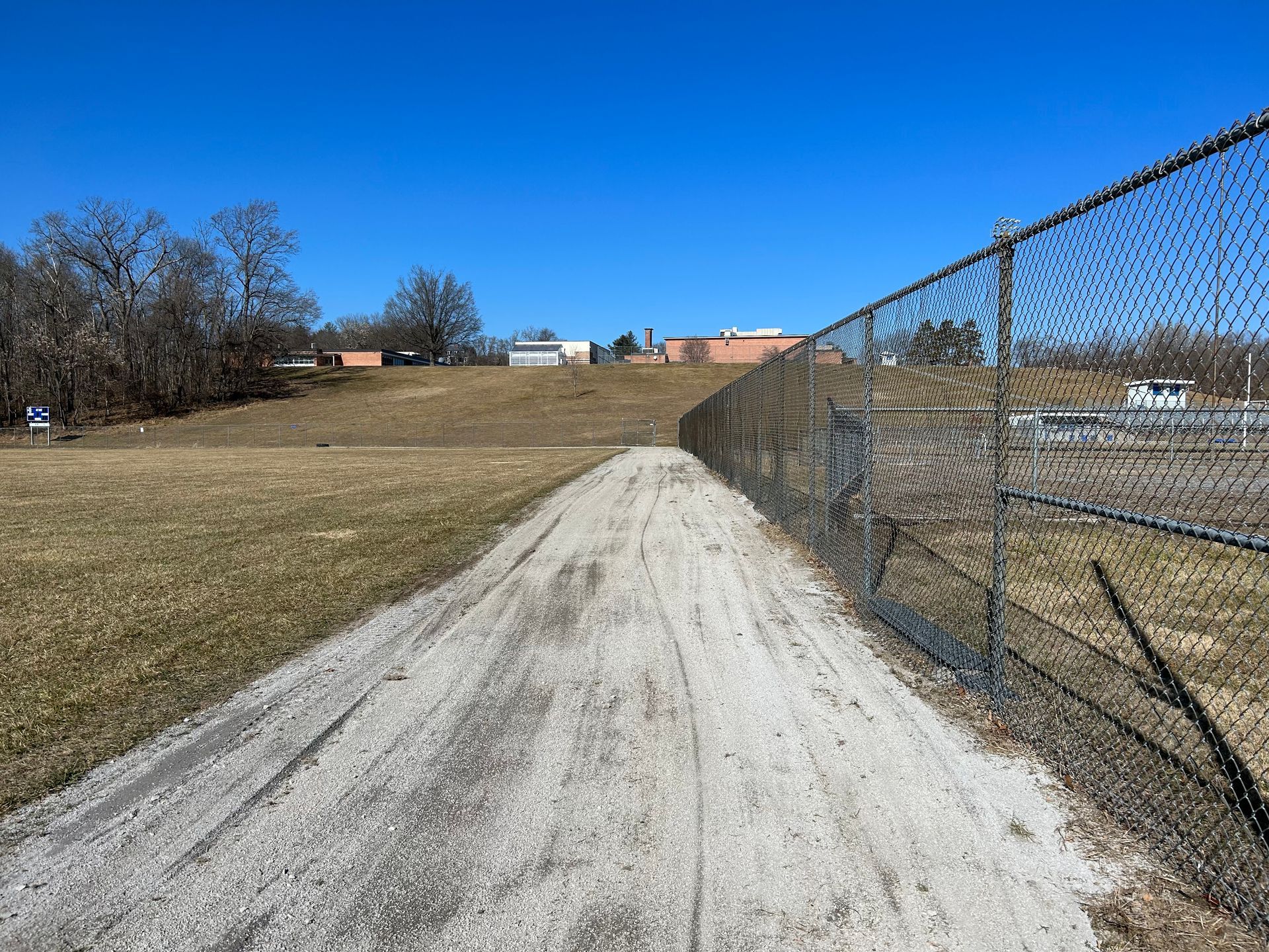 A dirt path runs alongside a chain-link fence under a bright blue sky, with a grassy hill and buildings in the distance.