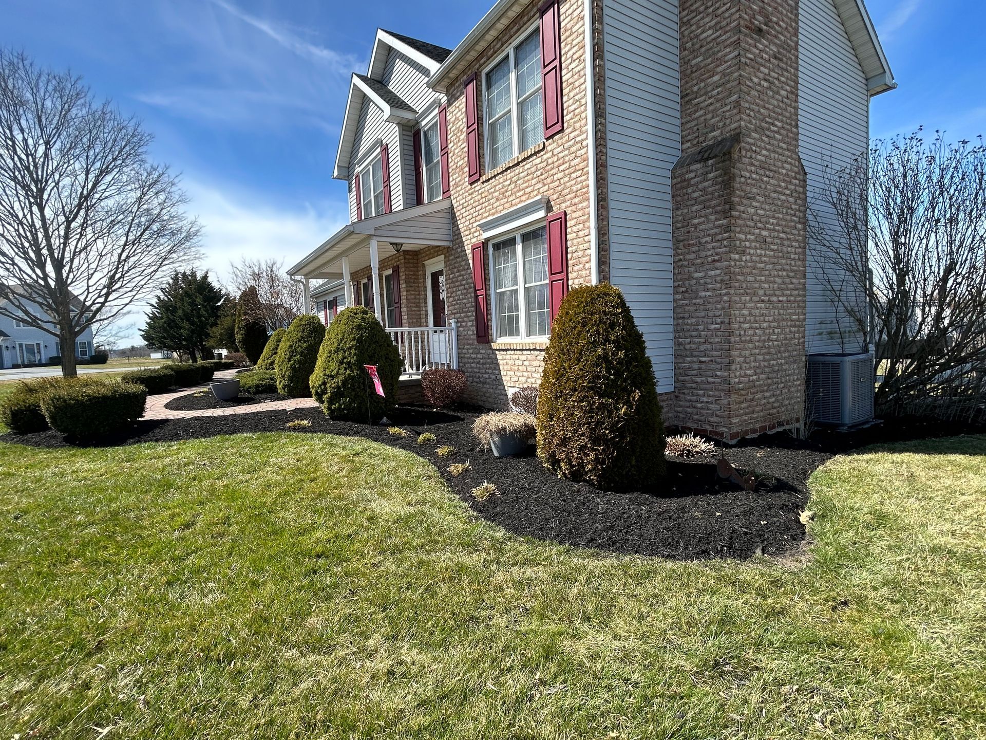 A two-story brick and siding house with dark mulch landscaping and trimmed evergreen shrubs under a clear blue sky.