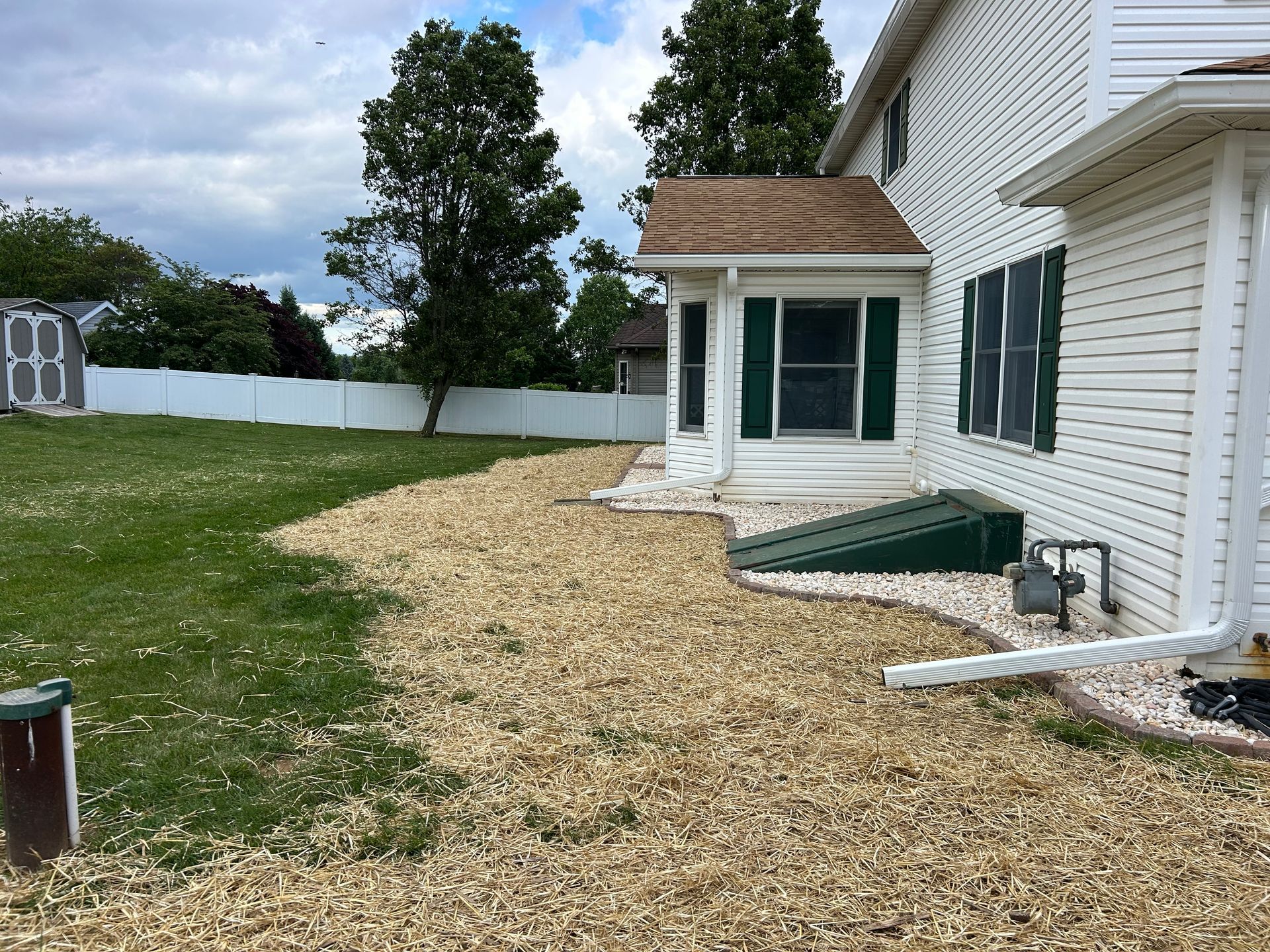 A side view of a white house with wood chips covering the ground, a green cellar door, and a white privacy fence.
