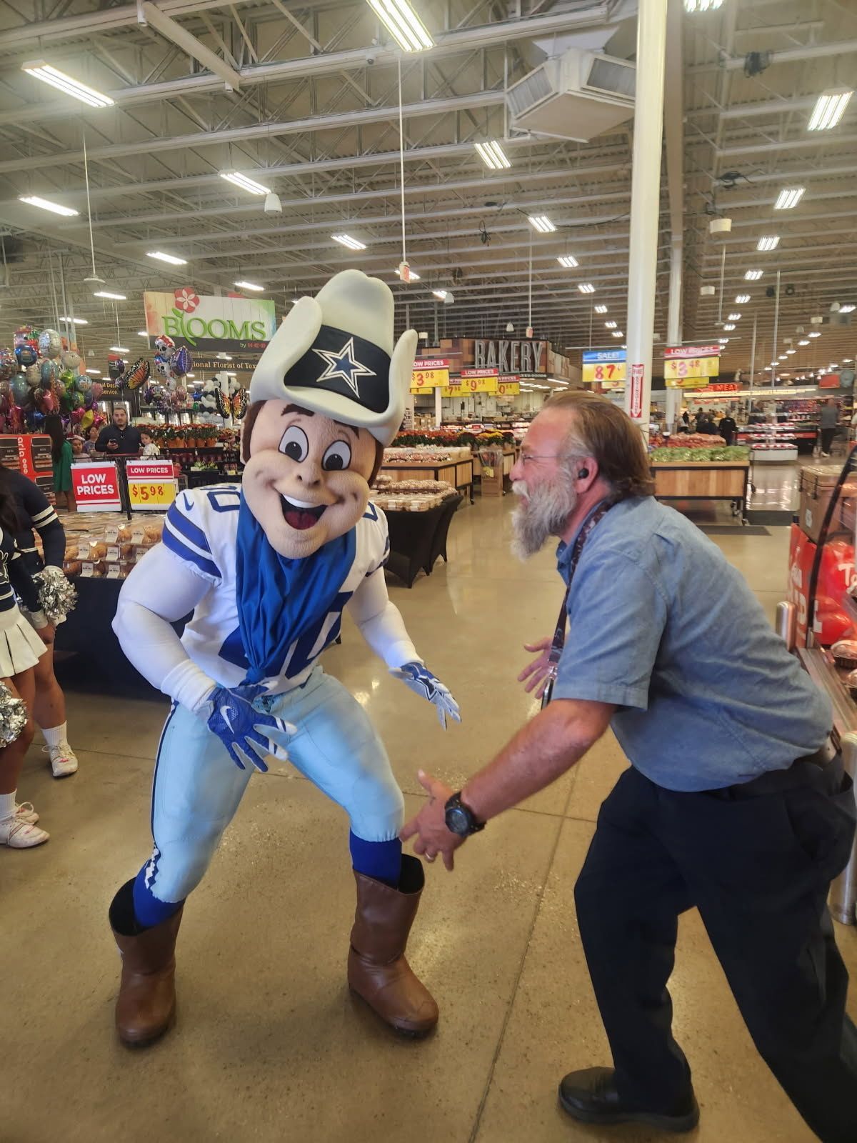 A Dallas Cowboys mascot in a hat and jersey poses with a man in a blue button-down shirt inside a brightly lit grocery store.