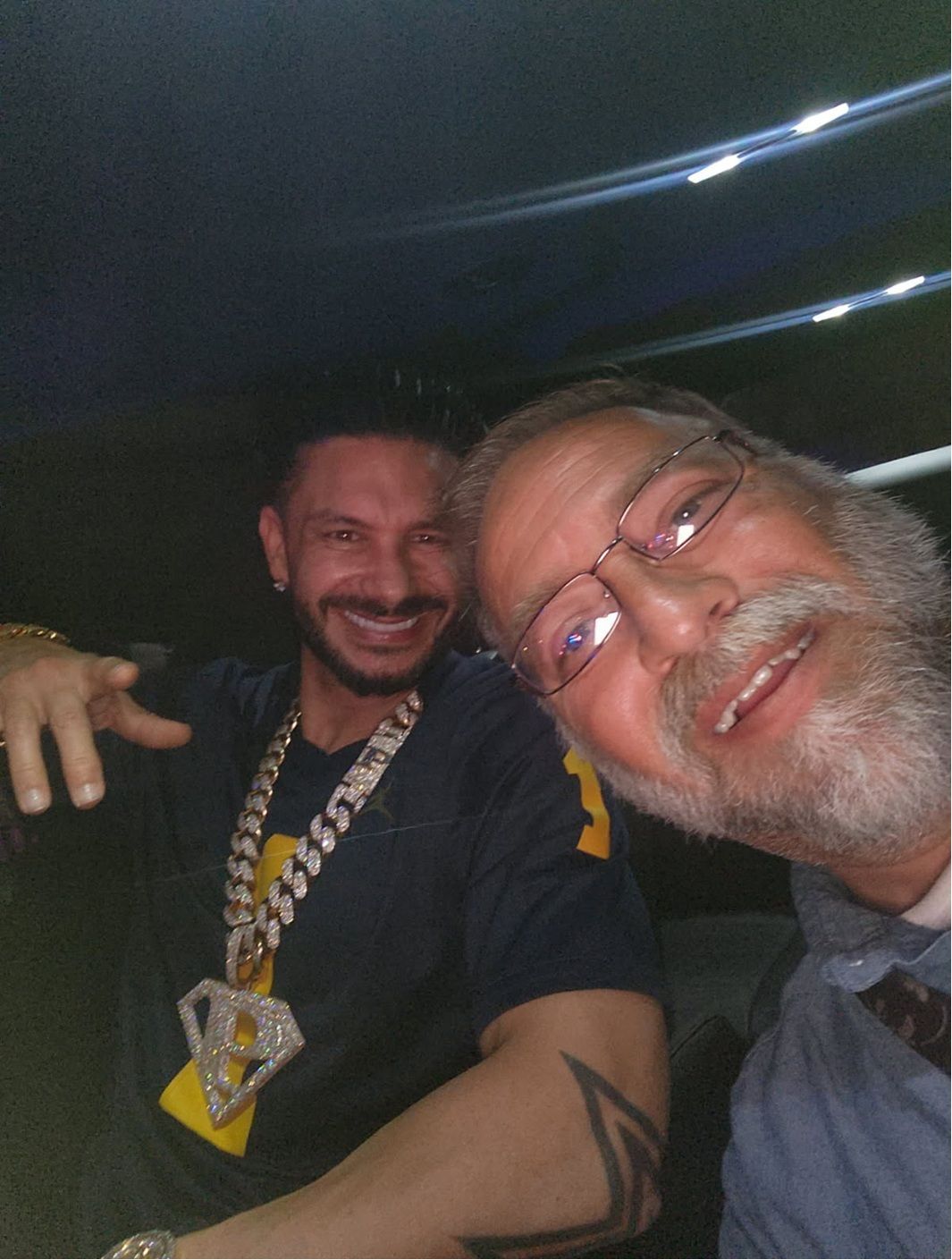 A man with a large diamond-encrusted chain smiles next to a man with a gray beard and glasses in a dimly lit setting.