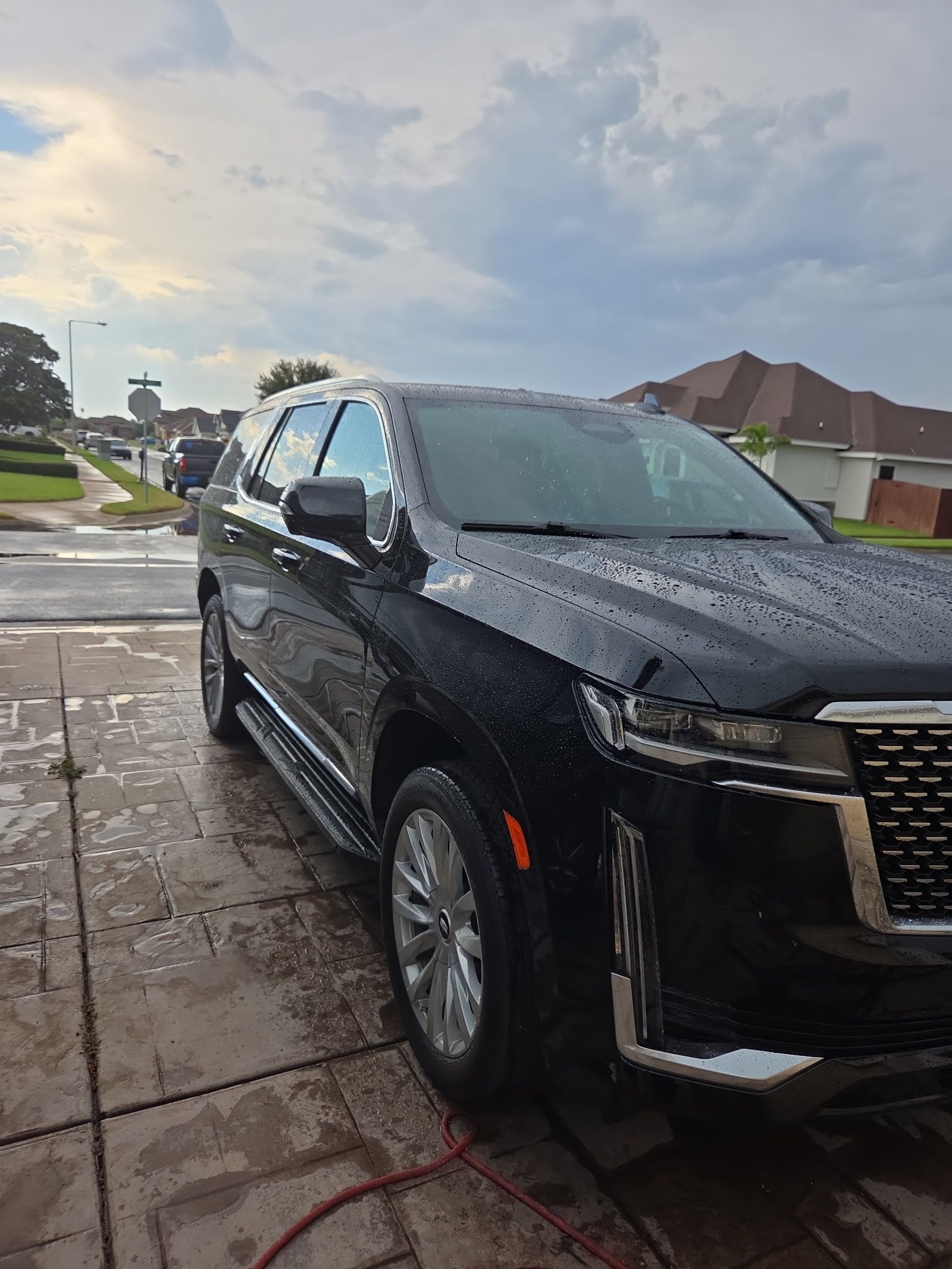 A black Cadillac Escalade parked on a paved driveway with raindrops on its surface under a cloudy sky.