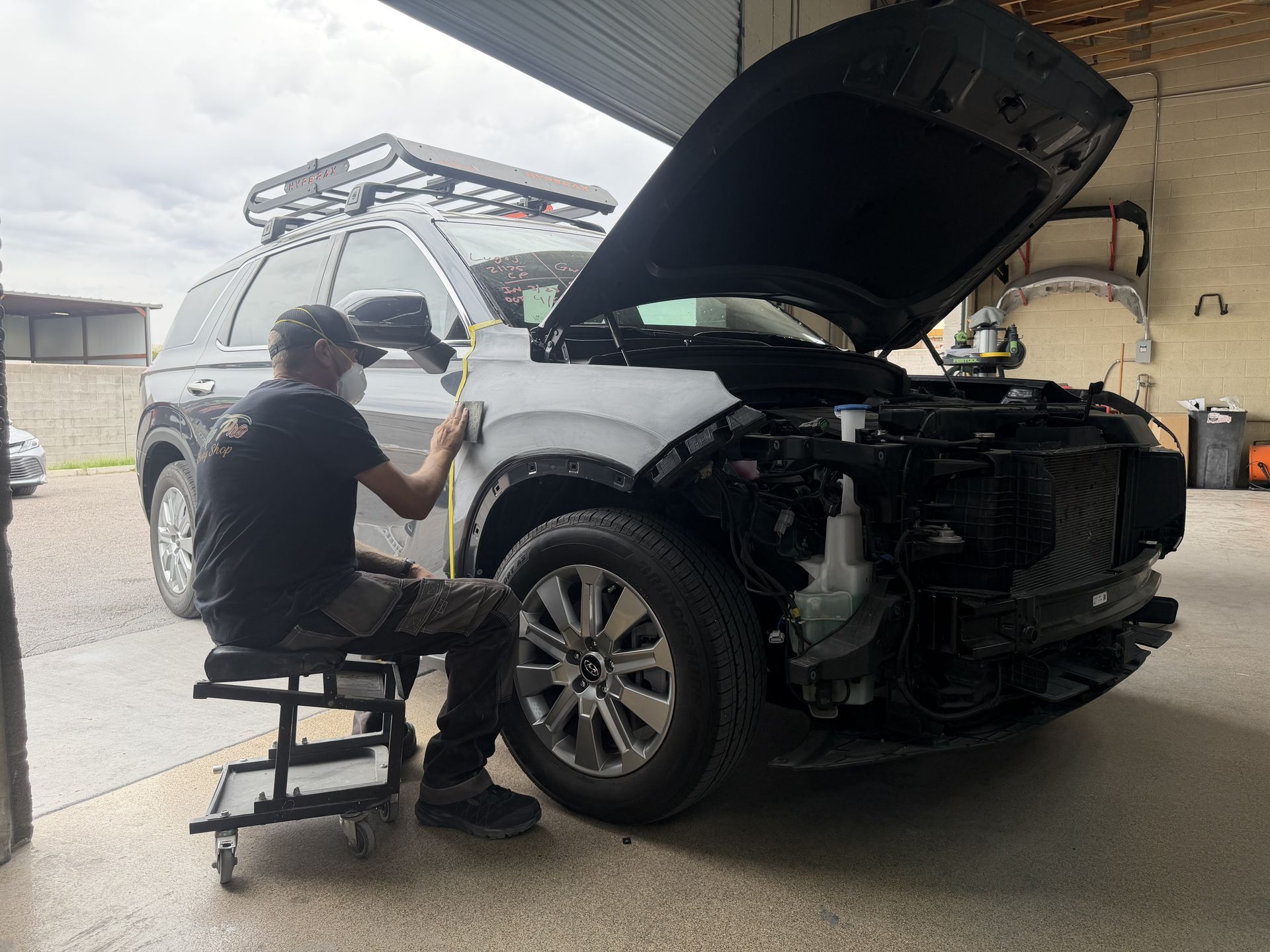 A person wearing a face mask sits on a rolling stool while sanding the front quarter panel of a white SUV in a shop.