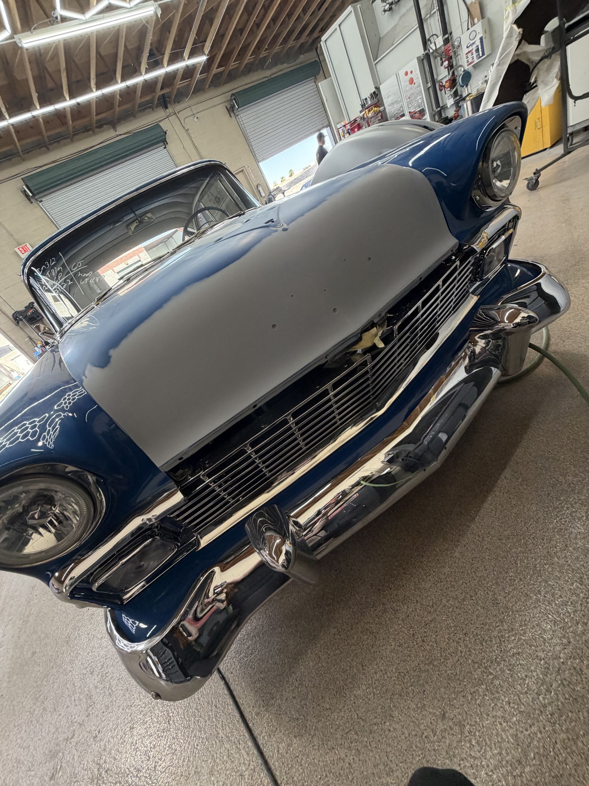 A blue 1956 Chevrolet with a gray primer hood sits inside a garage.