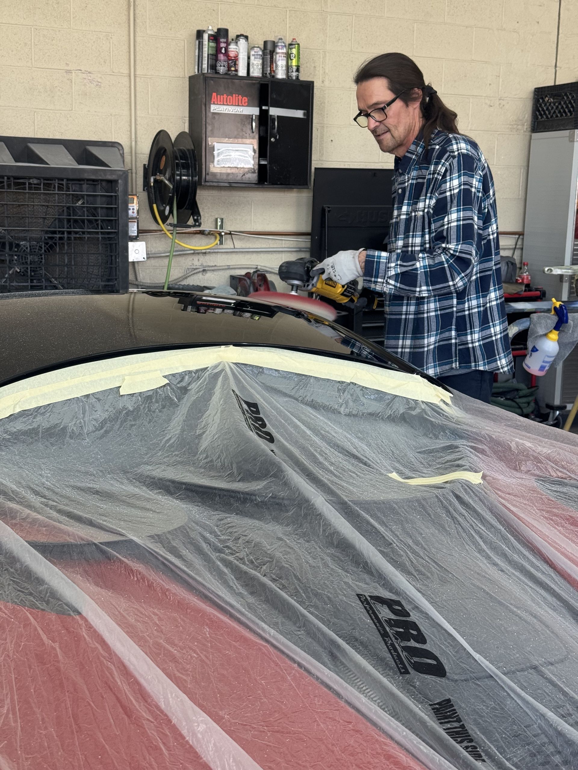 A person in a plaid shirt uses a tool on a car covered in plastic sheeting inside a garage.