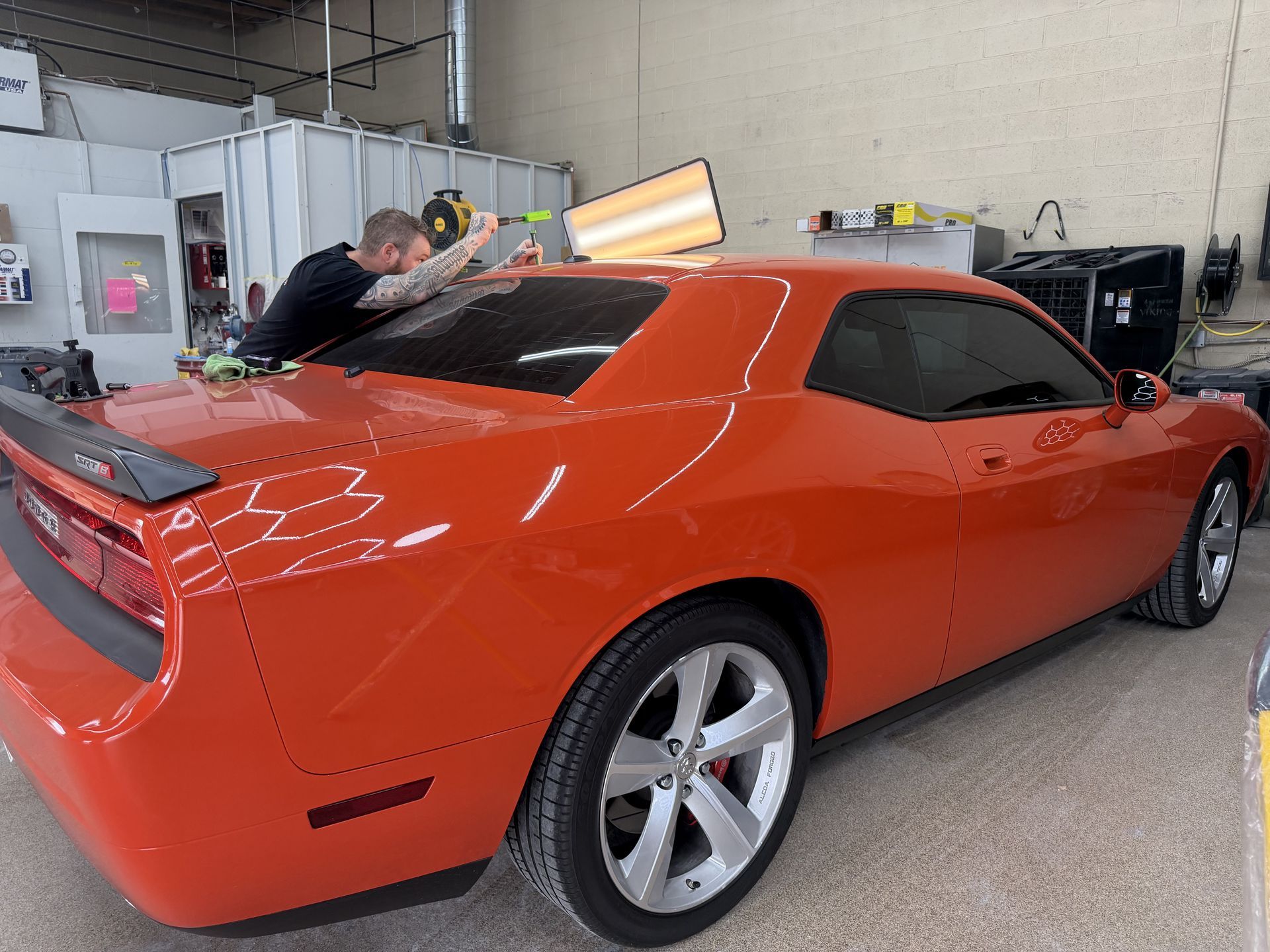 A person using a tool to perform paintless dent repair on the roof of an orange Dodge Challenger in a garage.