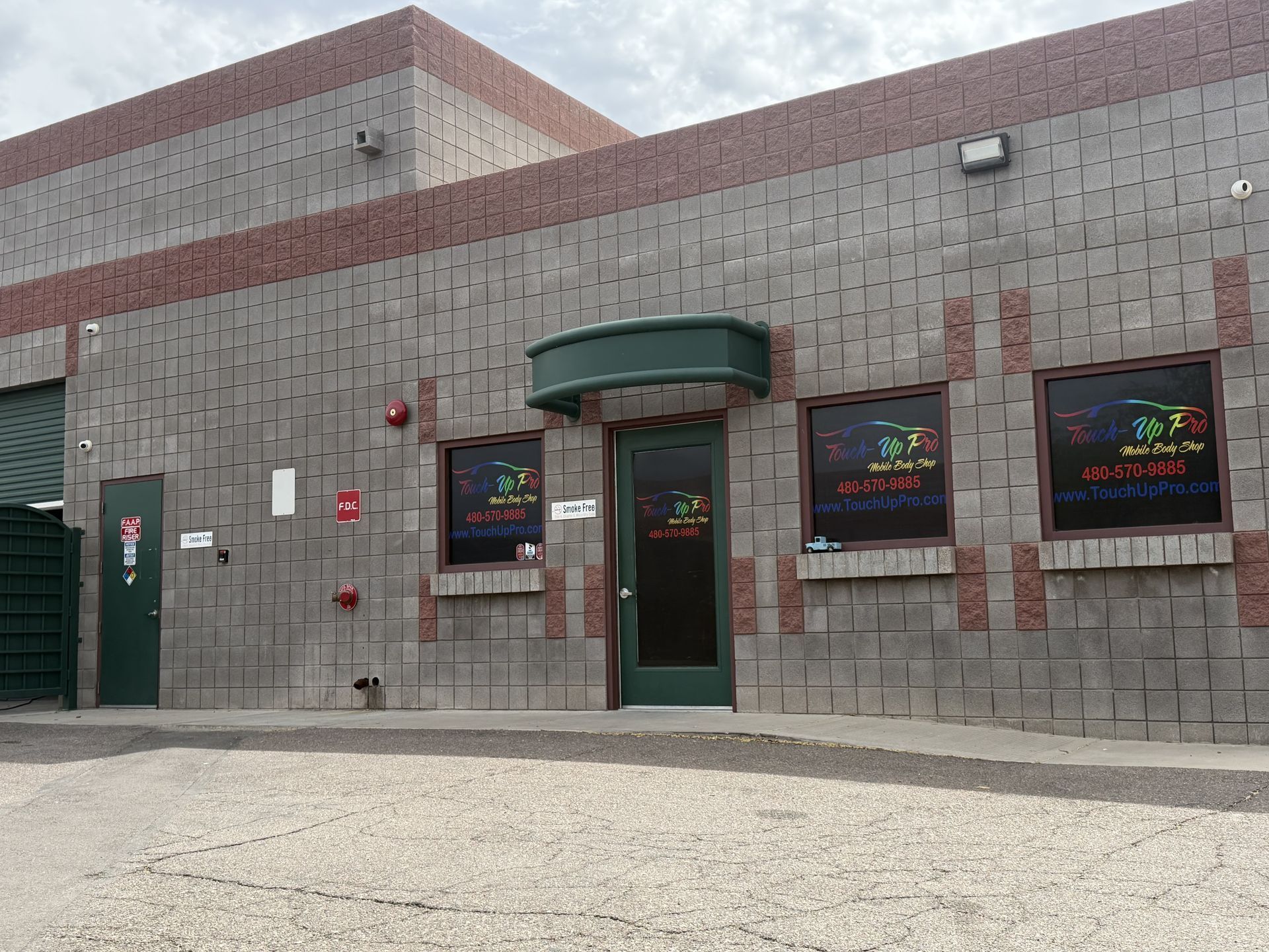 A grey and red brick building exterior featuring a central door with a green awning and two windows with colorful signs.