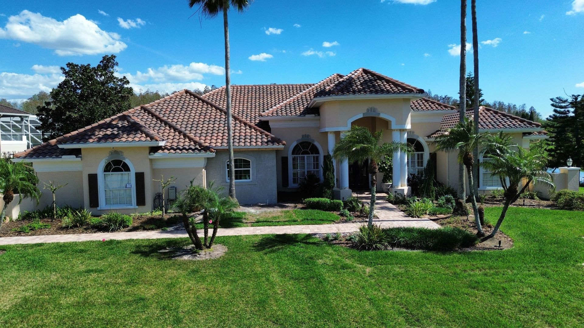 Single-story tan stucco home with a terracotta tiled roof, palm trees, and a green lawn under a bright blue sky.