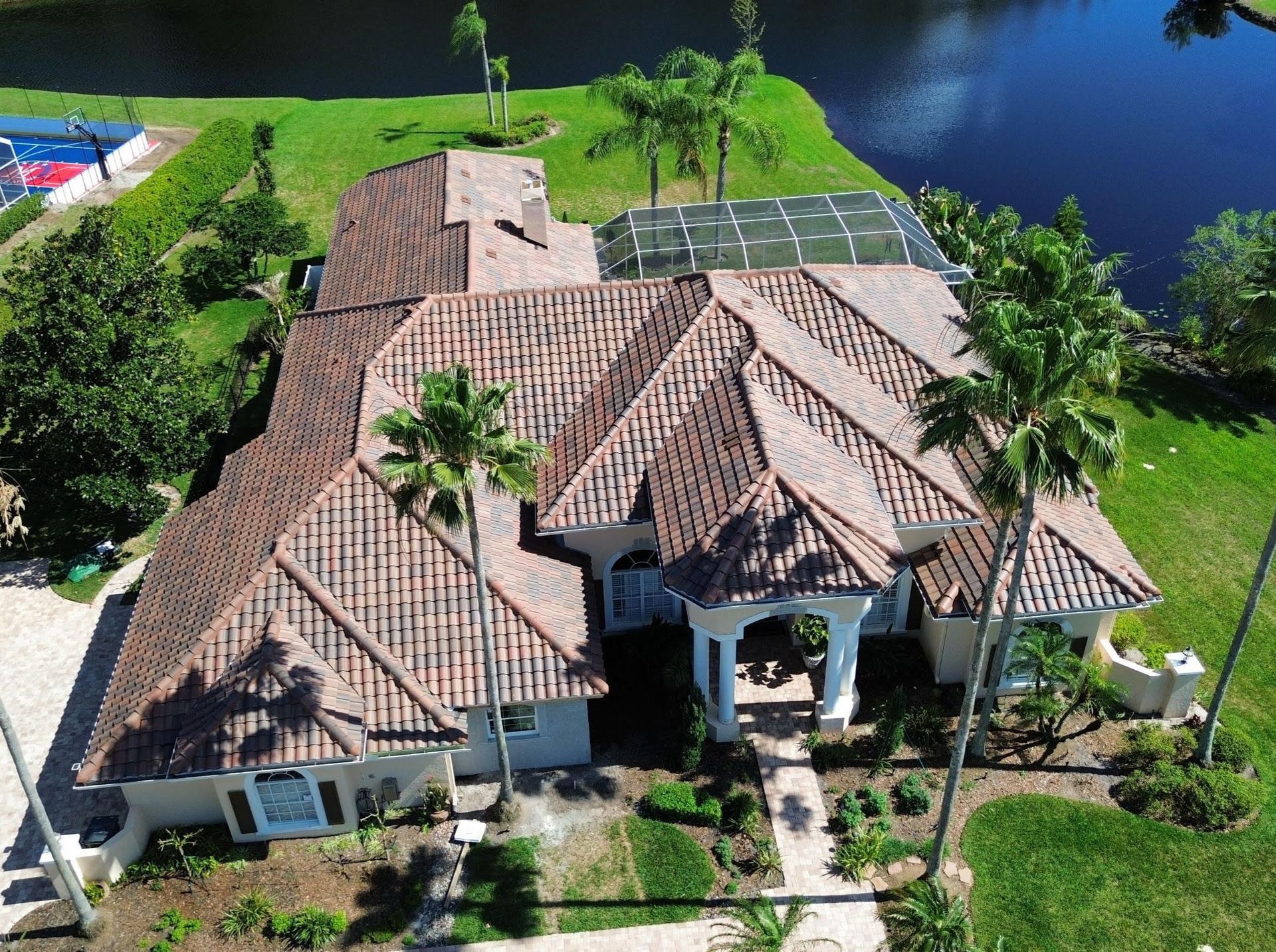 An aerial view of a single-story house with a brown tile roof, surrounded by a lawn, palm trees, and a backyard pond.