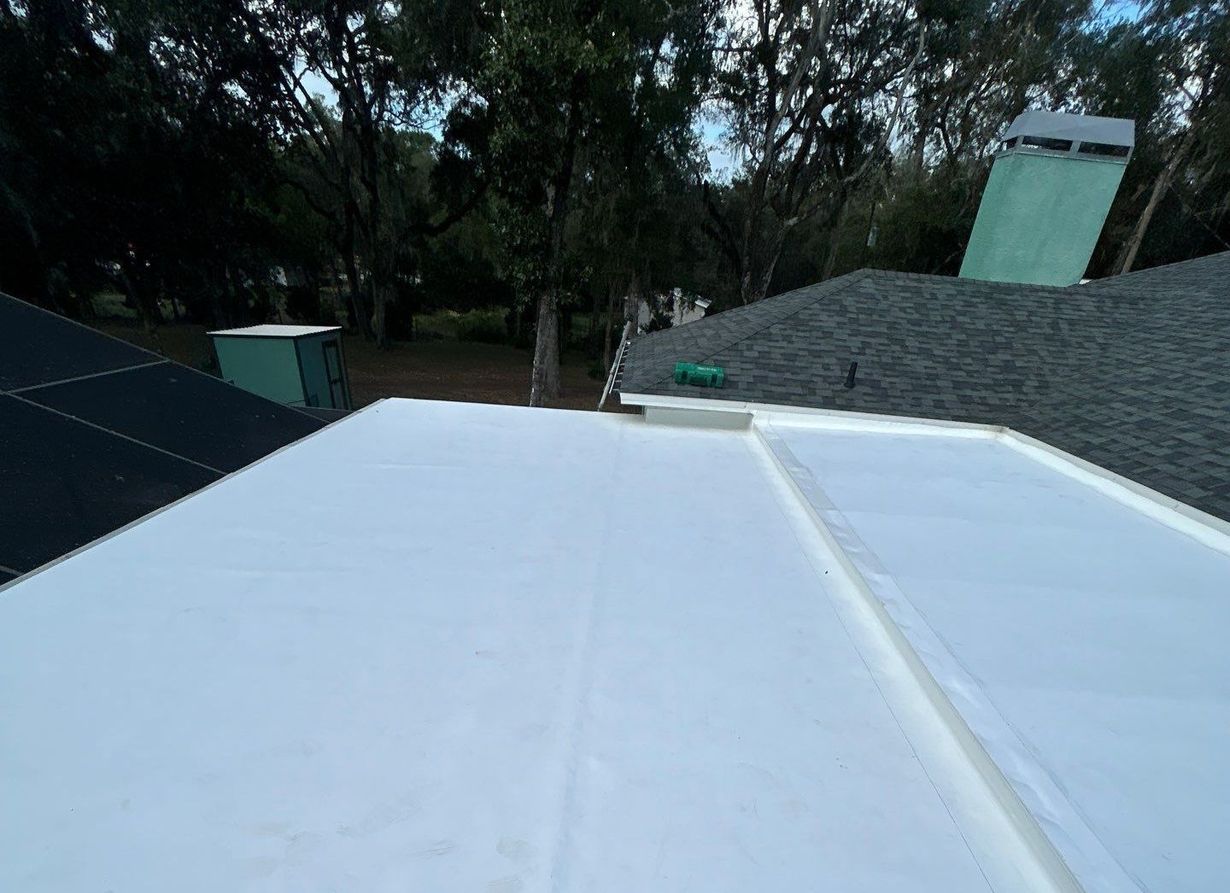A white flat roof with adjacent shingled roof, trees, and a green chimney in the background.