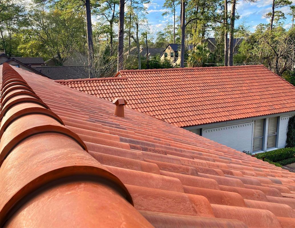 Red tile roof on a house in a wooded area, with a small vent and trees in the background.