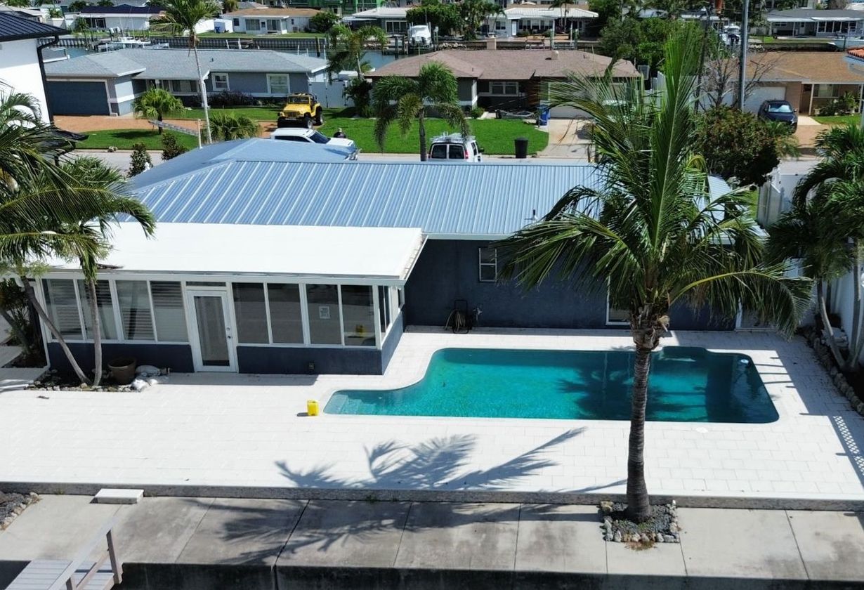 House with a blue roof, pool, and palm trees. Located near a waterfront, on a sunny day.