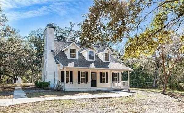 White house with black shutters, dormer windows, and a porch, set in a yard with trees.