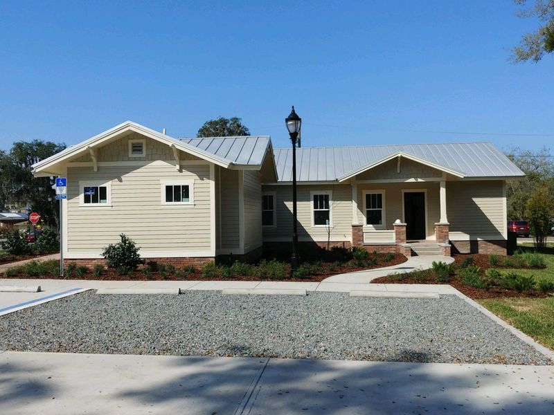 Tan building with a silver roof and dark brick accents; gravel parking area.
