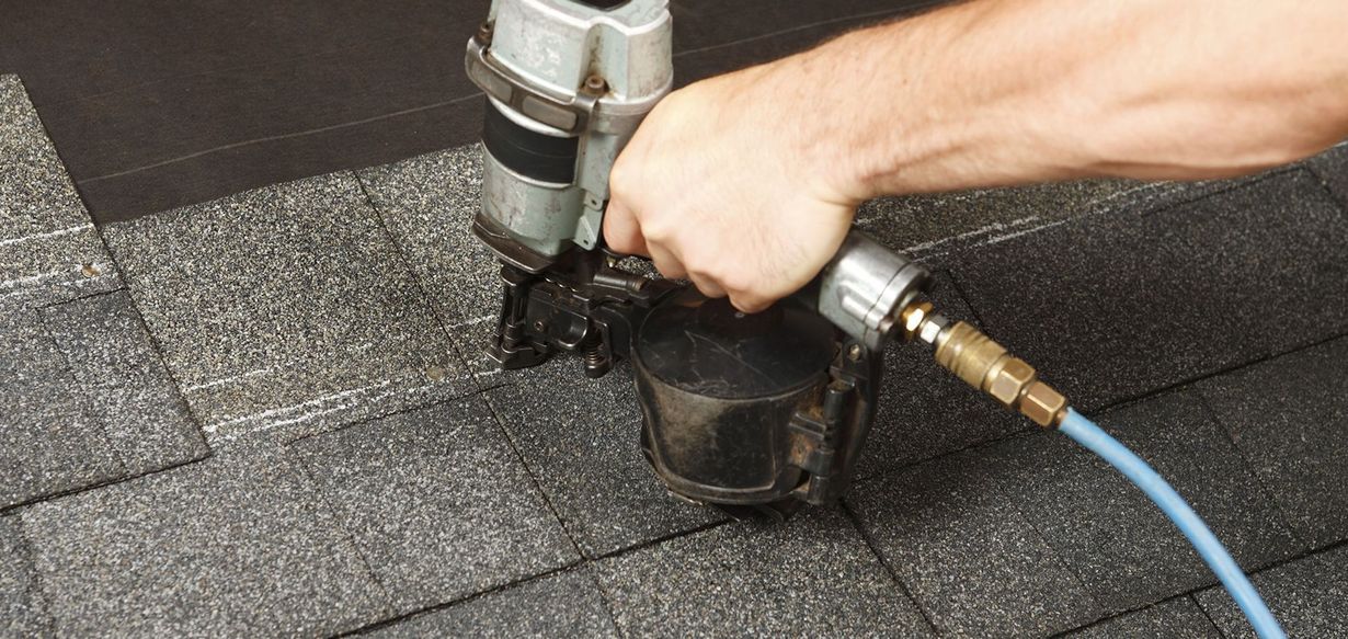 A hand uses a nail gun to install asphalt shingles on a roof.