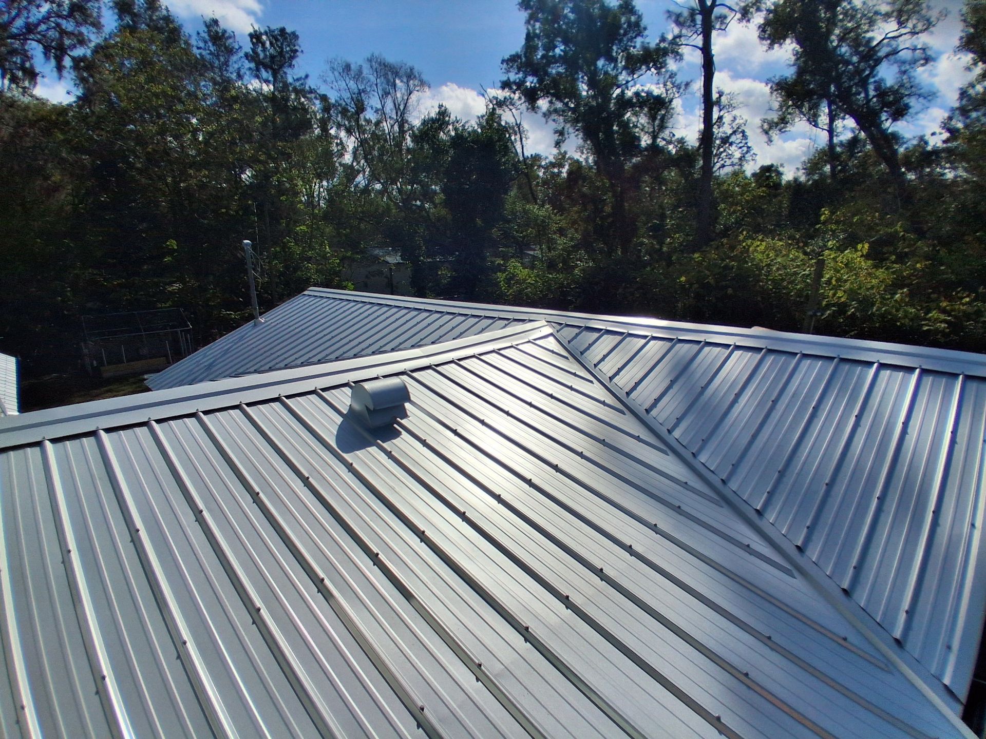 Silver metal roof with ridges, vents, and treeline background.