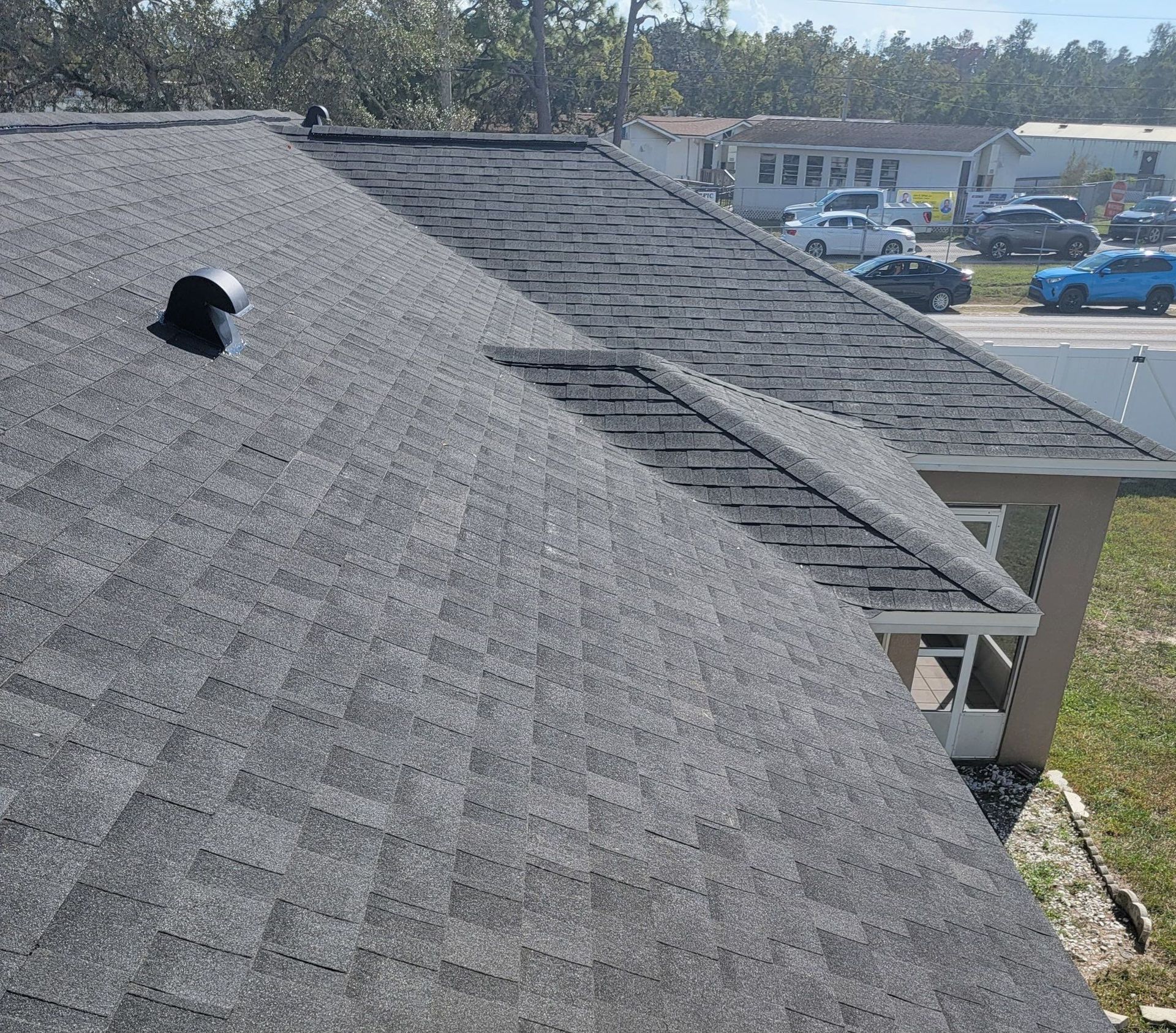 Gray shingle roof, chimney, and angled roof edge. Residential setting with parked cars in the background.